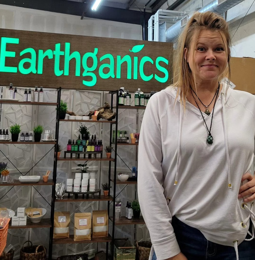 Woman with blonde hair wearing a white hoodie and necklaces standing in front of Earthganics sign and shelves with products.