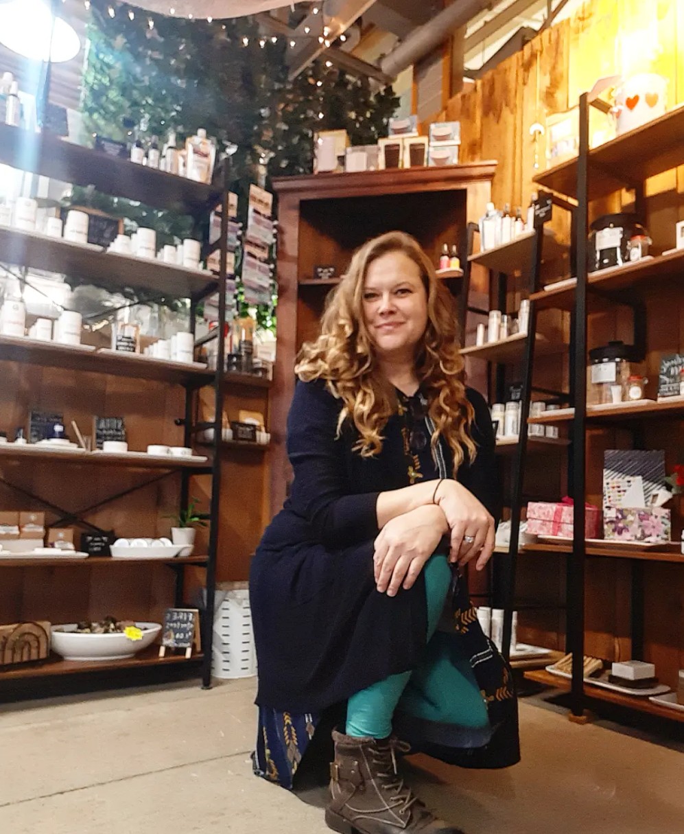Woman with long curly hair kneeling in store with shelves of products behind her.
