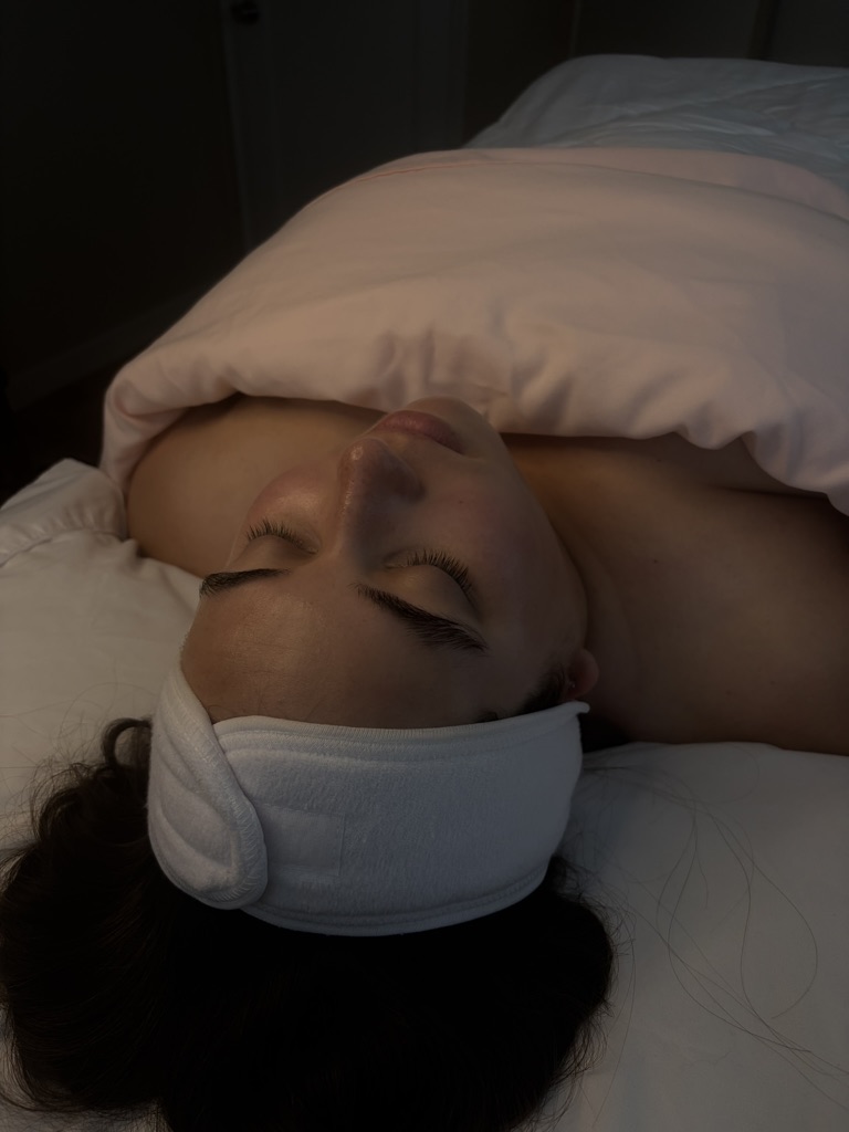 Person lying in bed with eyes closed, head resting on pillow, wearing a headband, covered partially by blanket.