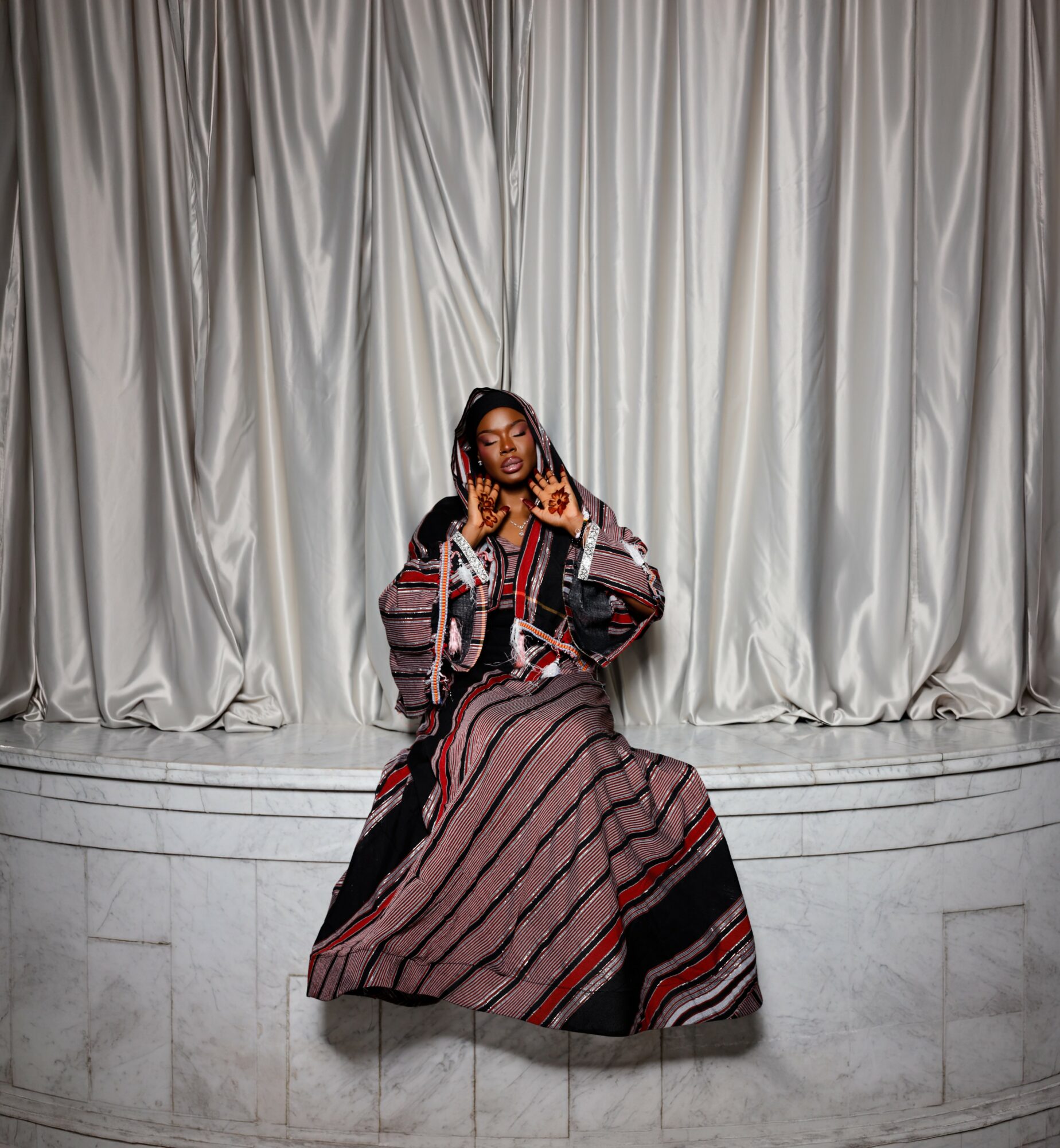 Person sitting on stage with a curtain background, wearing a striped dress and jacket, holding a microphone, making a gesture.