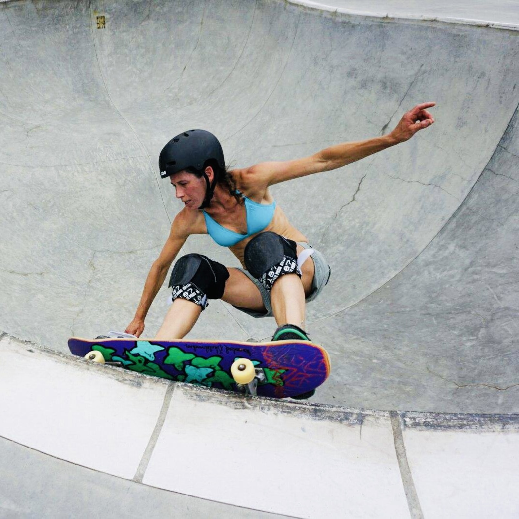 Person skateboarding in a skate park wearing a helmet and knee pads, crouched low on the board, with arms extended for balance.