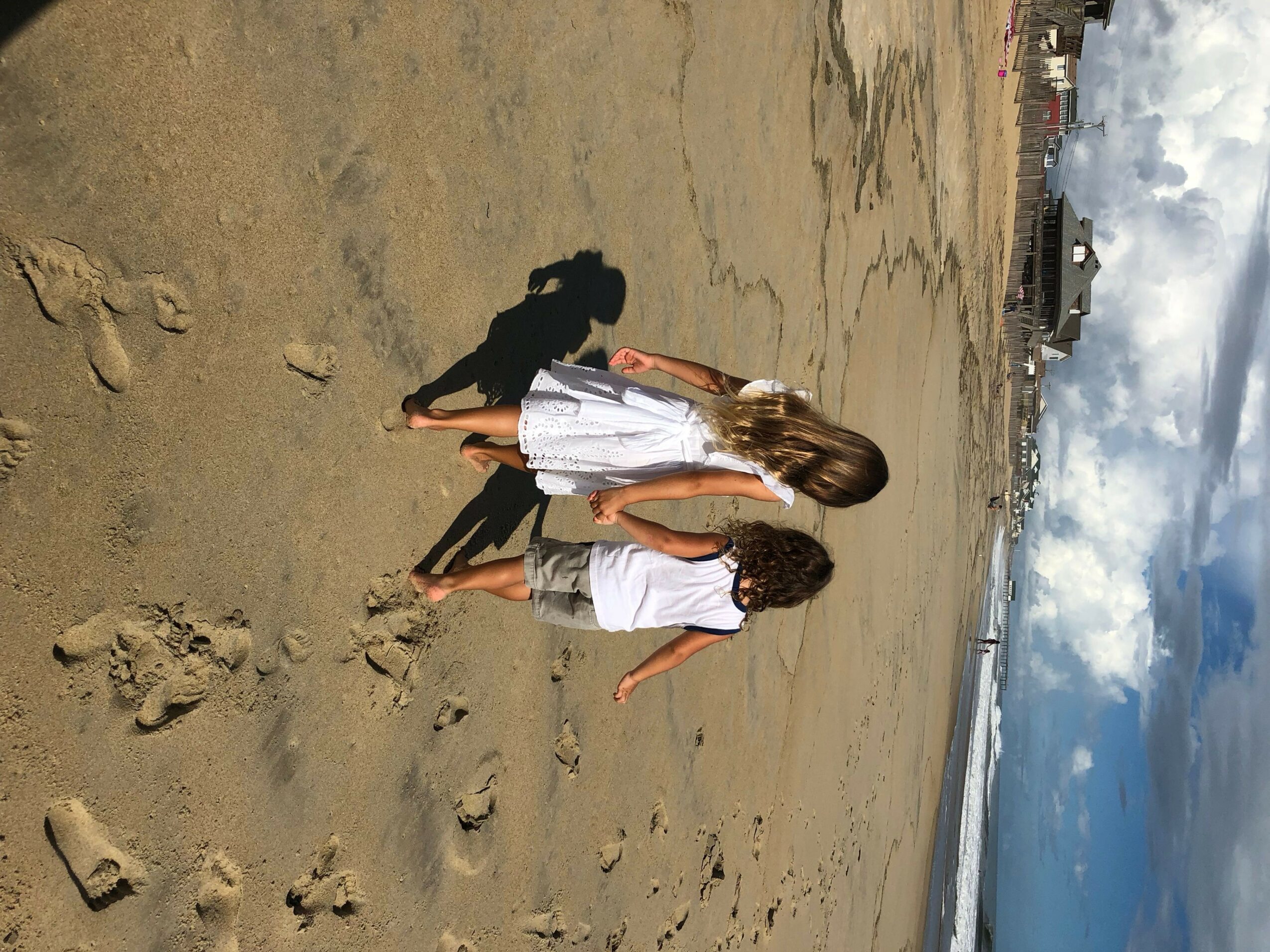 Two children walk on sandy beach with footprints, ocean, and sky in background, under partly cloudy sky.