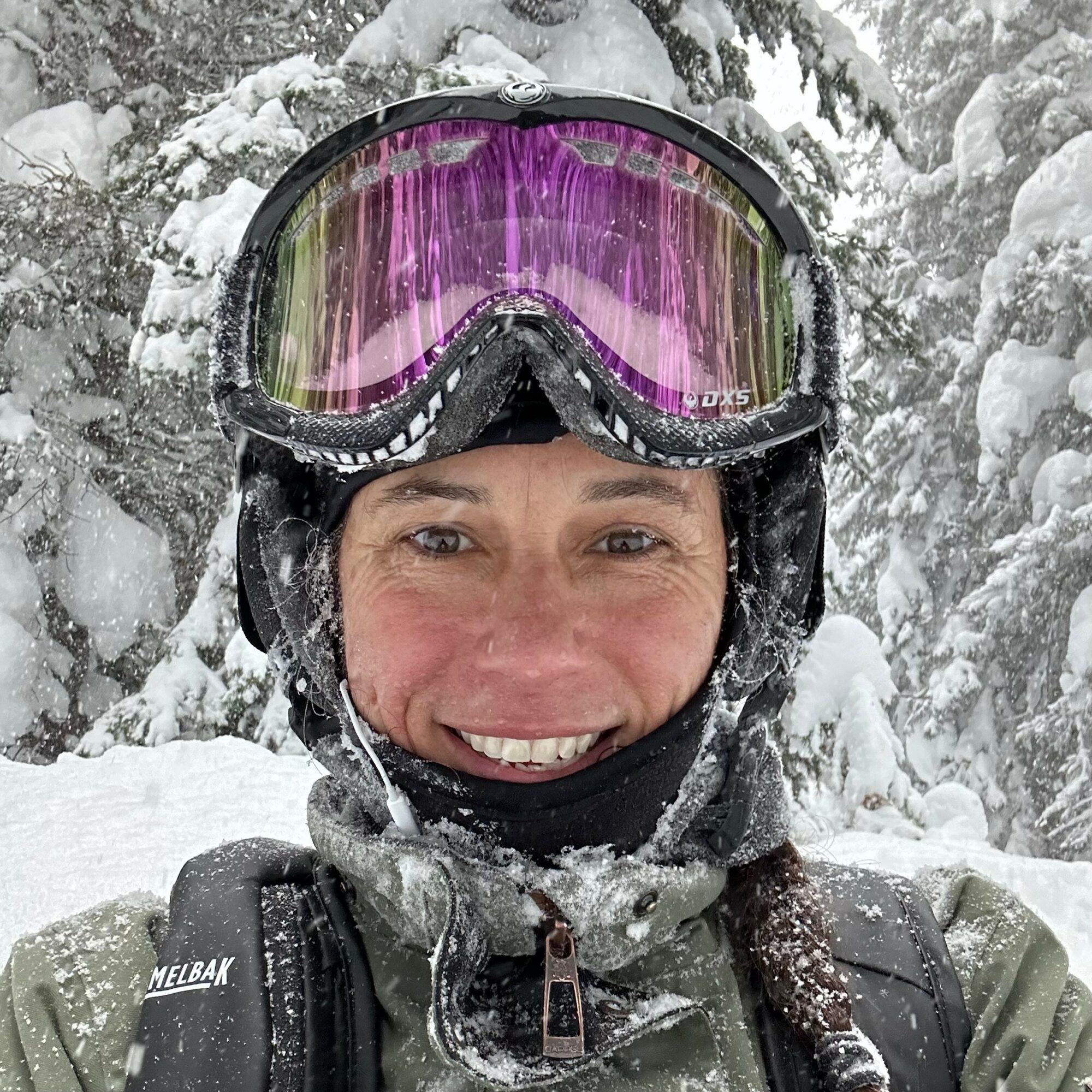 Person wearing ski goggles and winter gear smiling in snowy forest