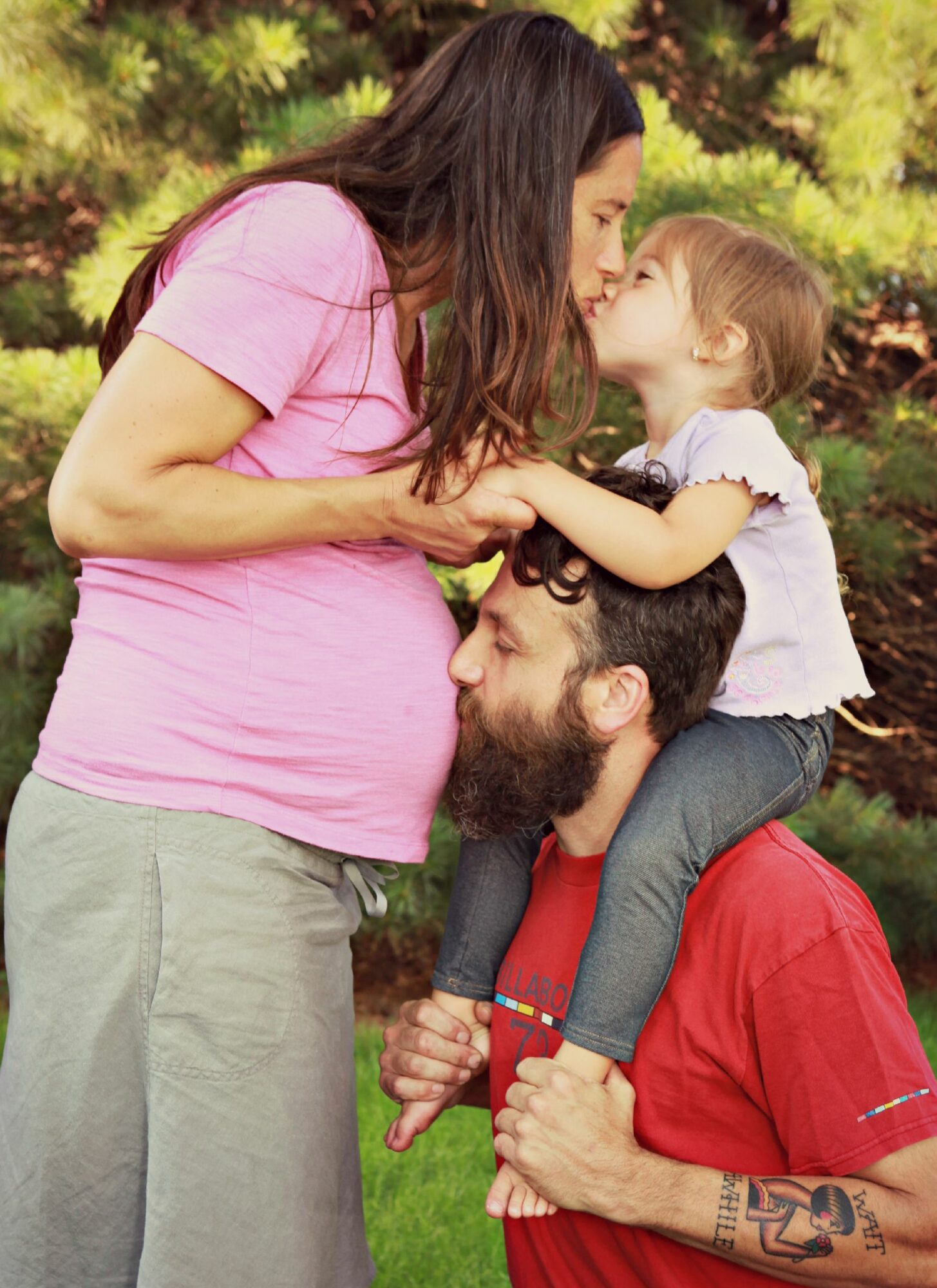 Woman and young girl touch noses outdoors, woman is pregnant, man supports girl on shoulders, trees in background.