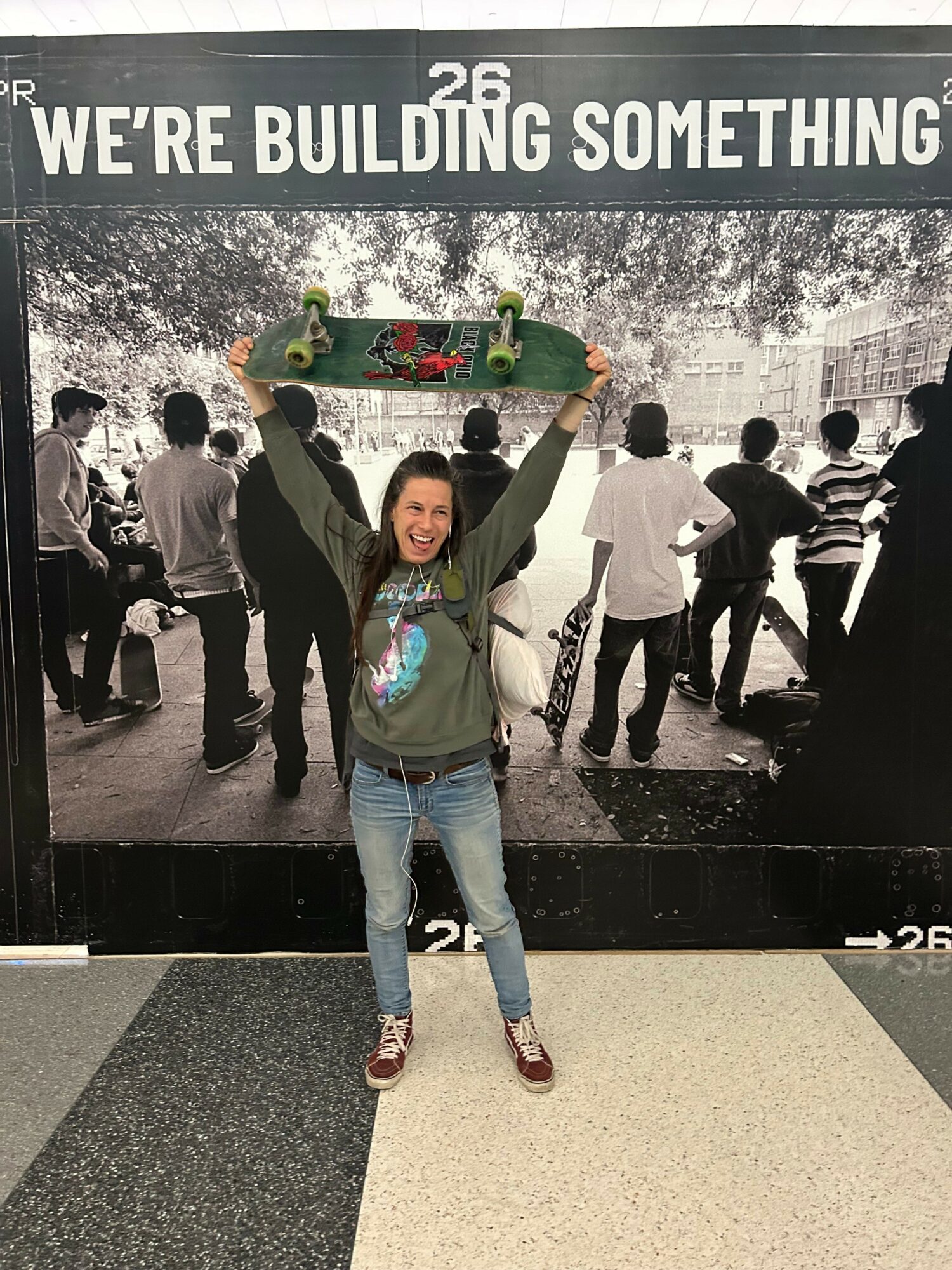 Person standing with arms outstretched in front of a group, black and white background, colorful clothing, and a sign reading 'WE'RE BUILDING SOMETHING'.
