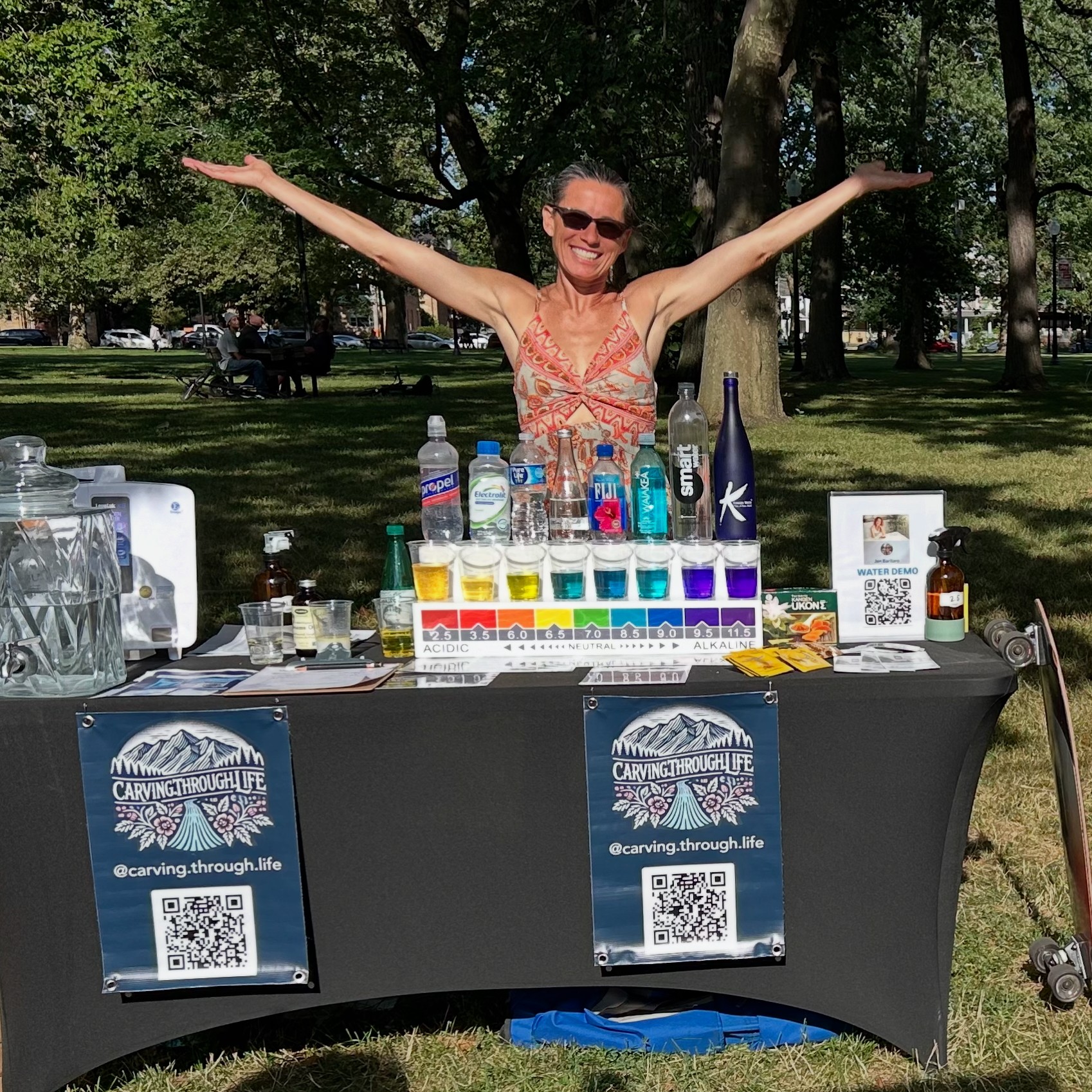 Woman with arms outstretched smiling behind a table with colorful drinks in a park.