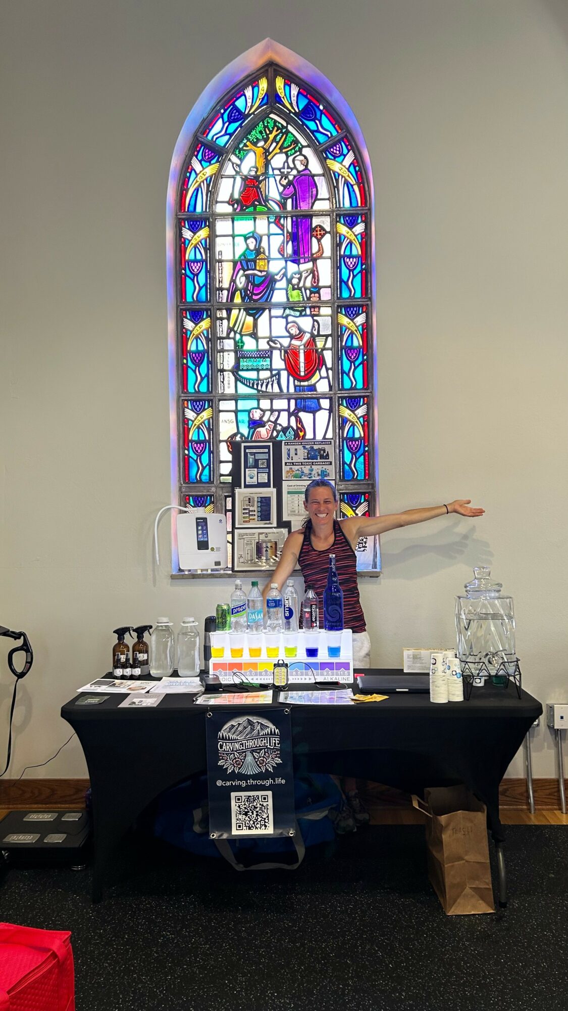 Person standing behind a table with colorful items in front of a stained glass window, arms outstretched, in a church setting.