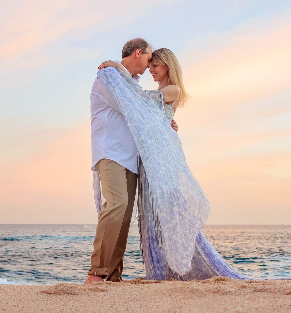 A couple stands close on a beach at sunset, embracing each other with the ocean in the background.