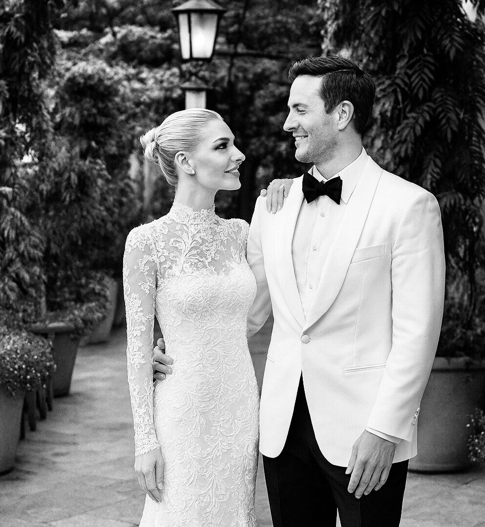 A couple in wedding attire standing outdoors, smiling at each other, surrounded by trees and potted plants.