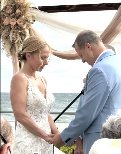 A bride and groom holding hands during a wedding ceremony outdoors near the ocean, with floral decorations and draped fabric.