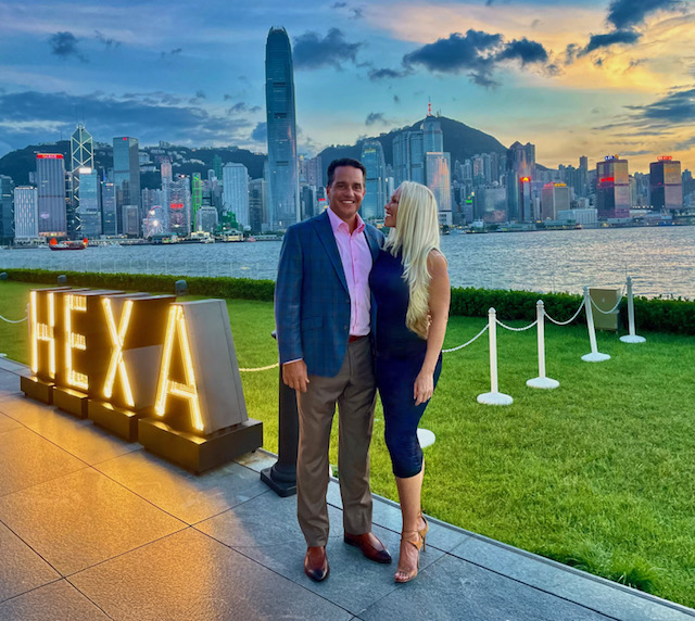 Two people stand near a waterfront with a city skyline and mountains in the background, illuminated sign reads 'HEXA'.