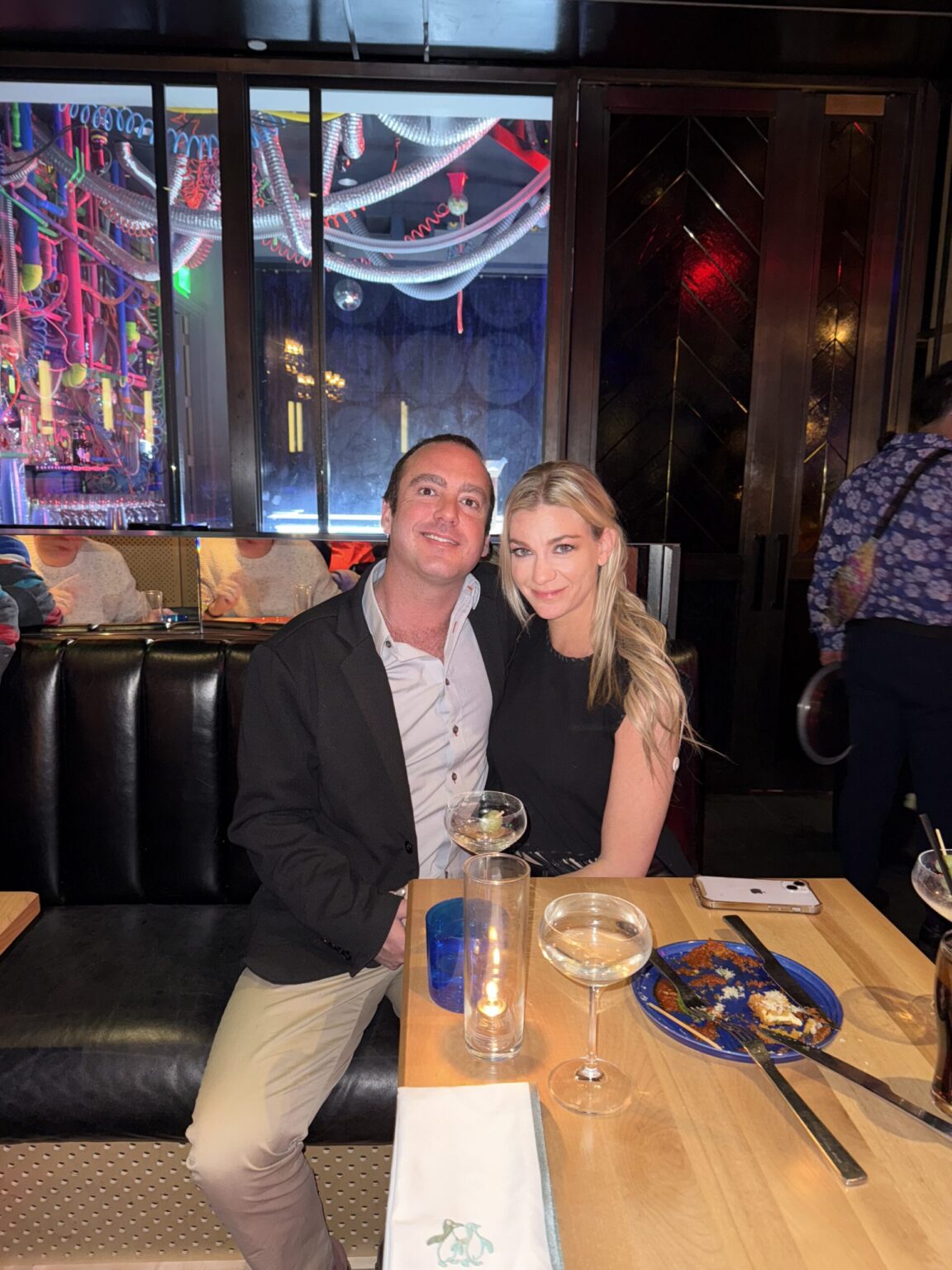 Two people sitting at a table in a restaurant, smiling, with colorful lights and decorations in the background.