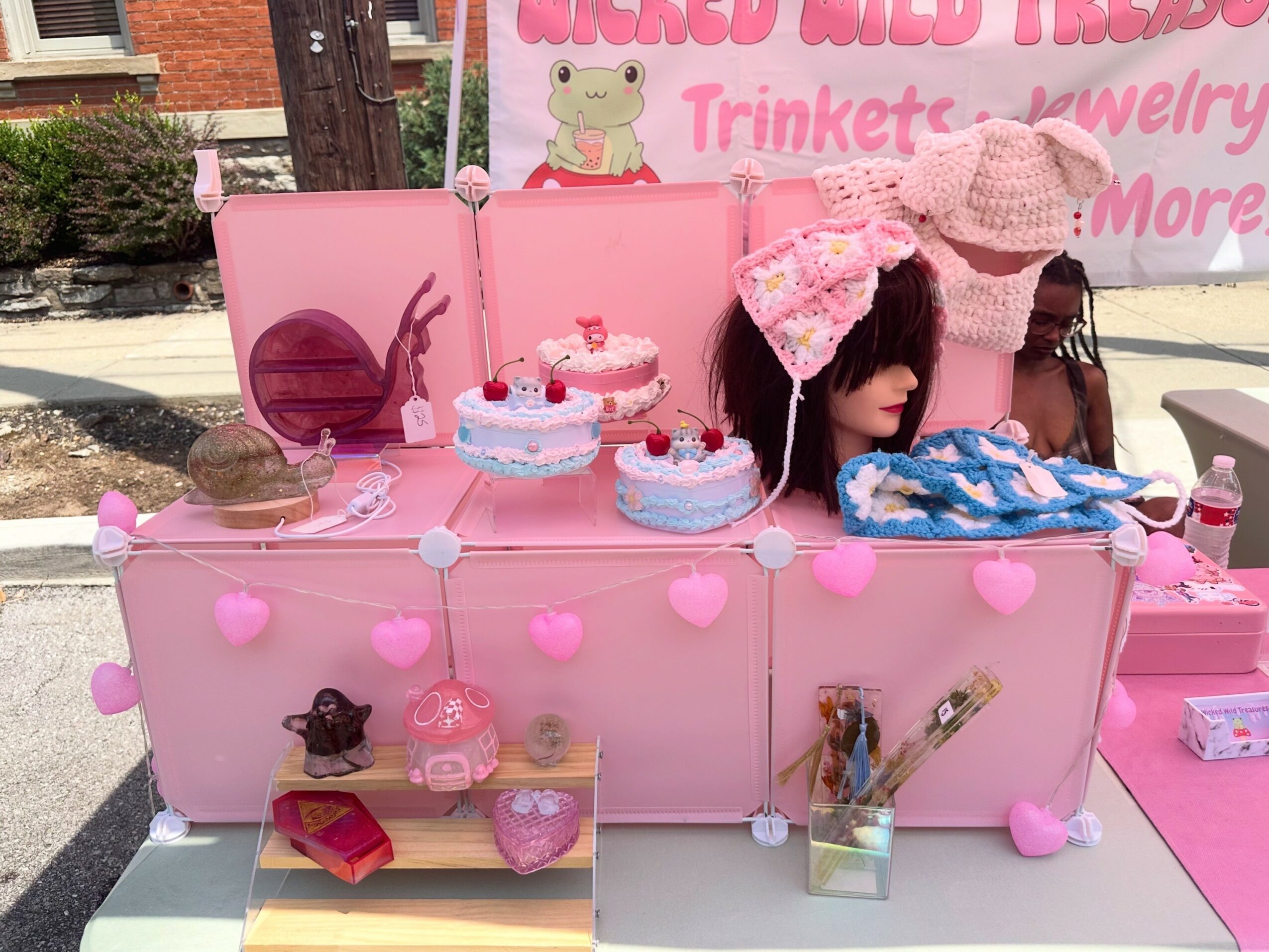 Pink display table with plush toys, cakes, and accessories, decorated with pink hearts and a mannequin head with a hat.