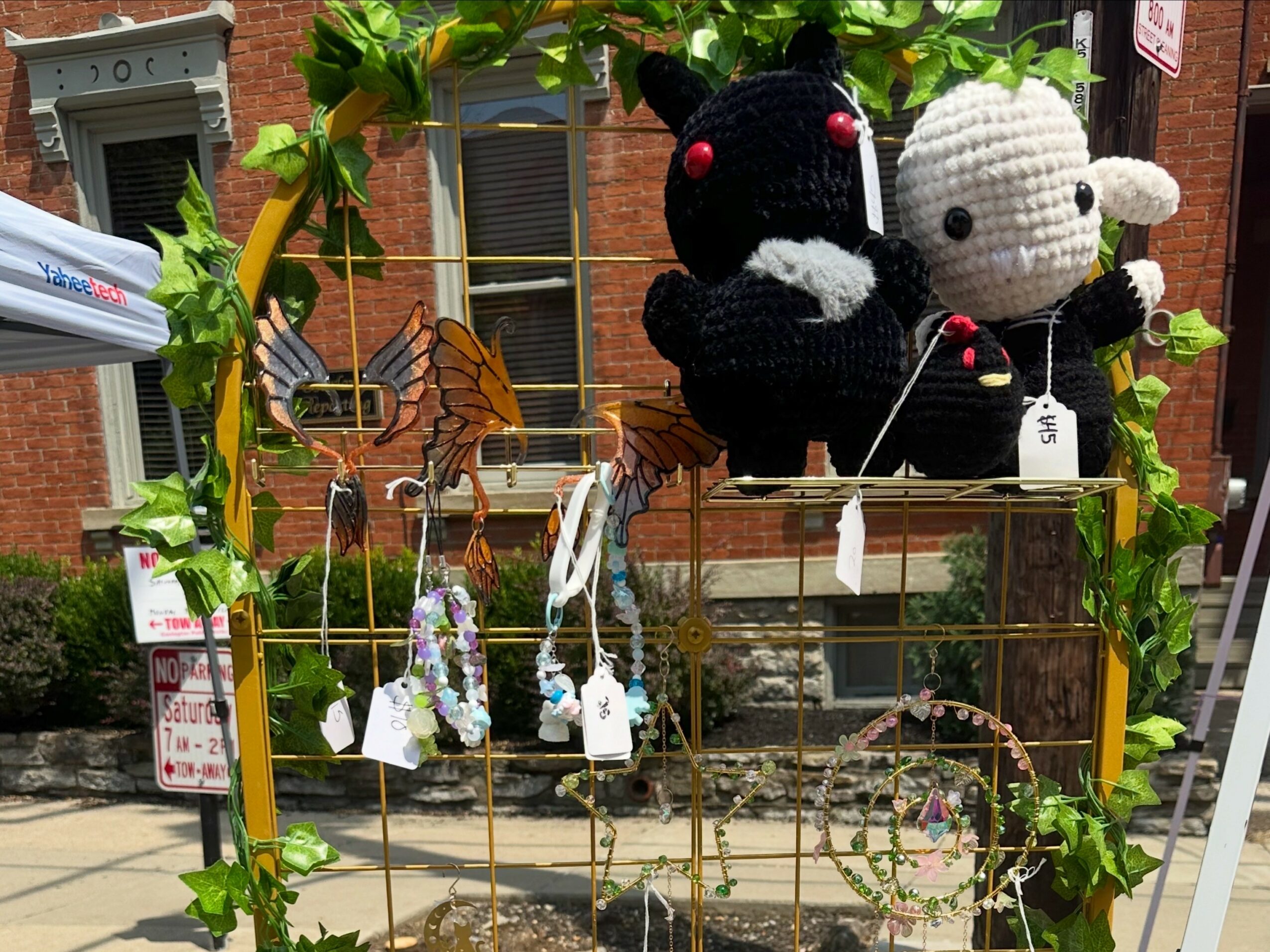Two plush toys, one black and one white, sit on a yellow metal shelf outdoors with plants and a brick building in background.