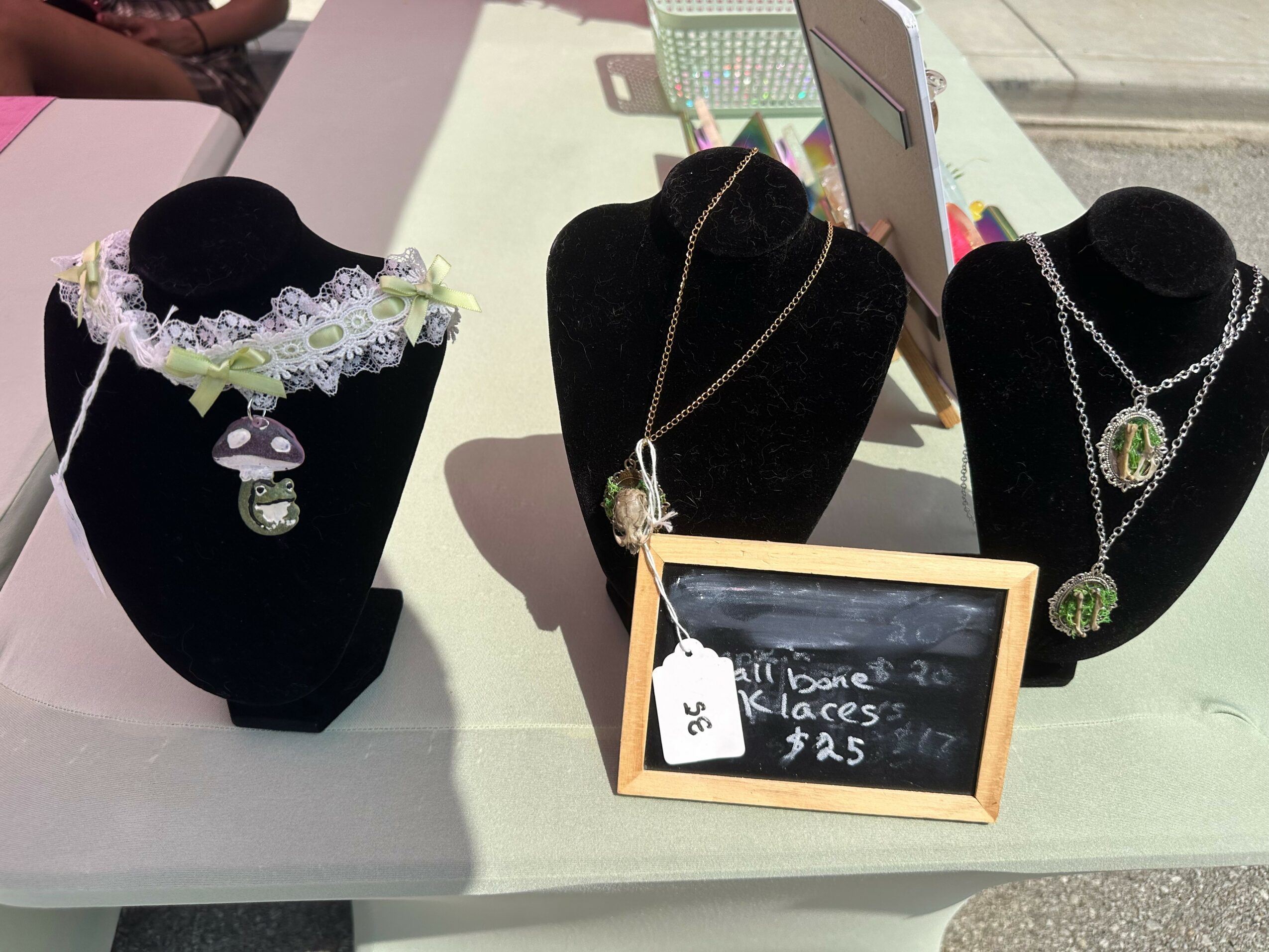 Three necklaces displayed on black busts, with a small chalkboard sign showing prices, outdoors on a table.