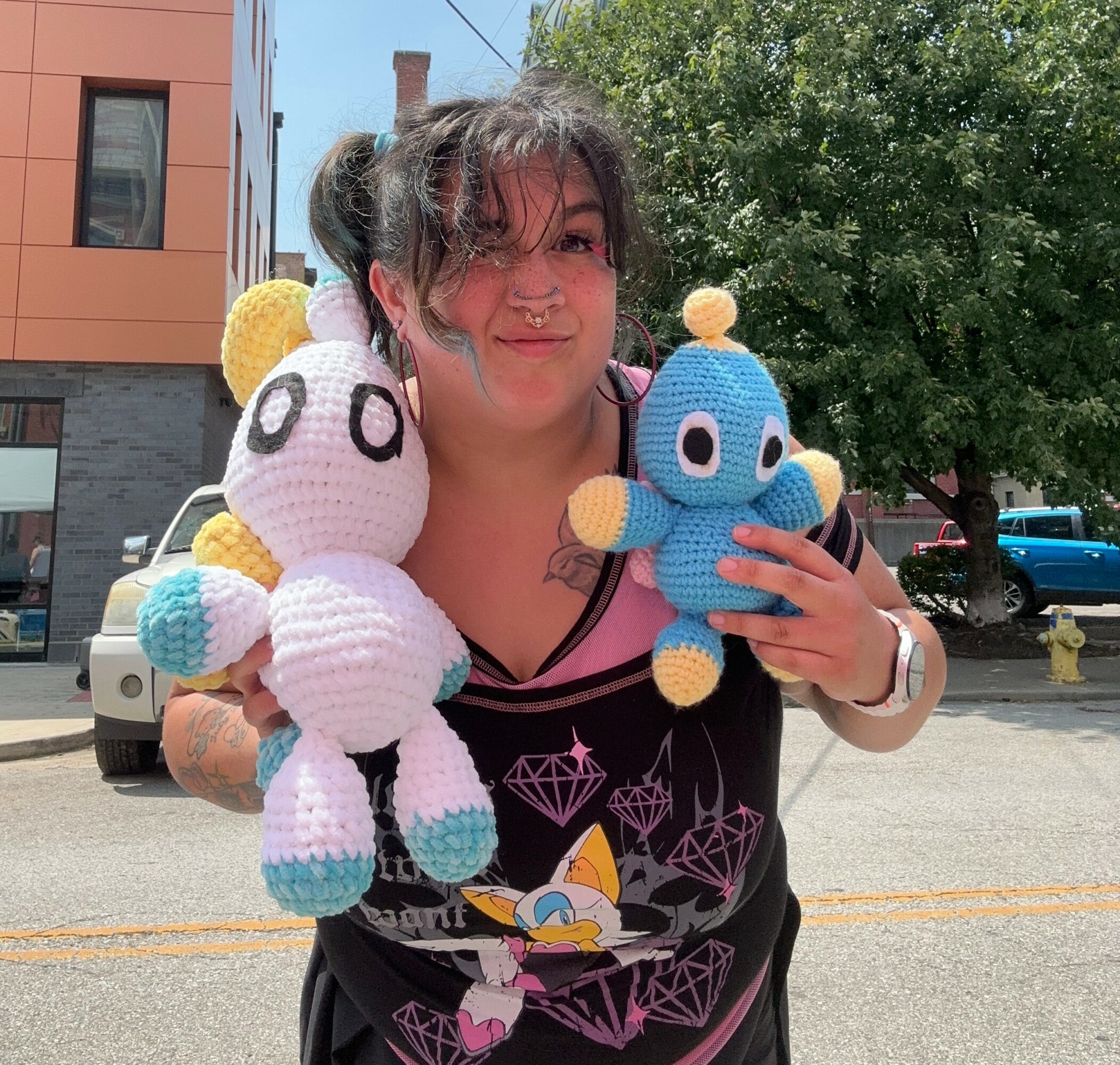 Person holding two plush toys, one white and one blue, outdoors on a sunny day with trees and buildings in background.