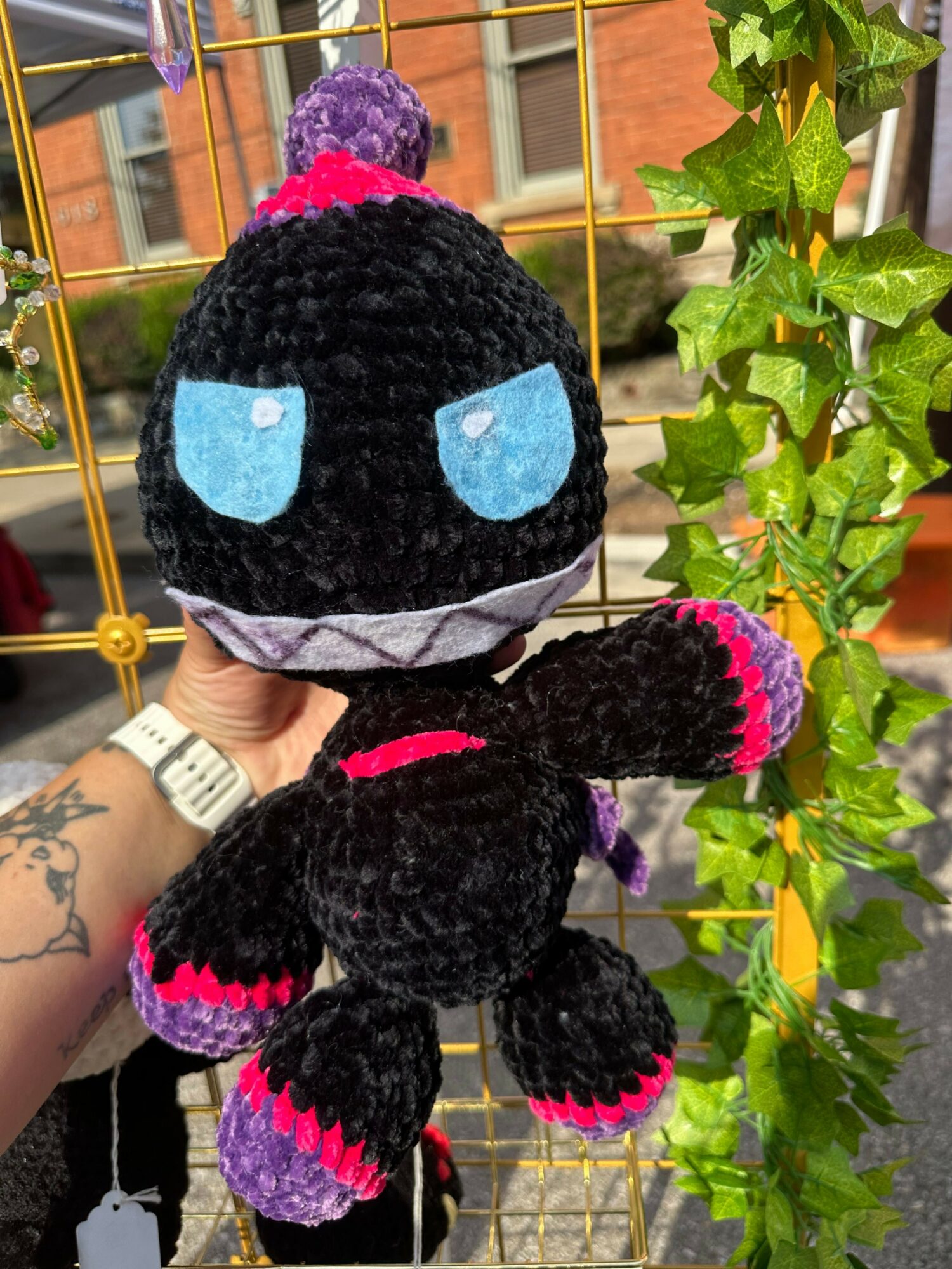 Person holding a black plush toy with blue eyes and pink accents, surrounded by green leaves and a yellow grid background.