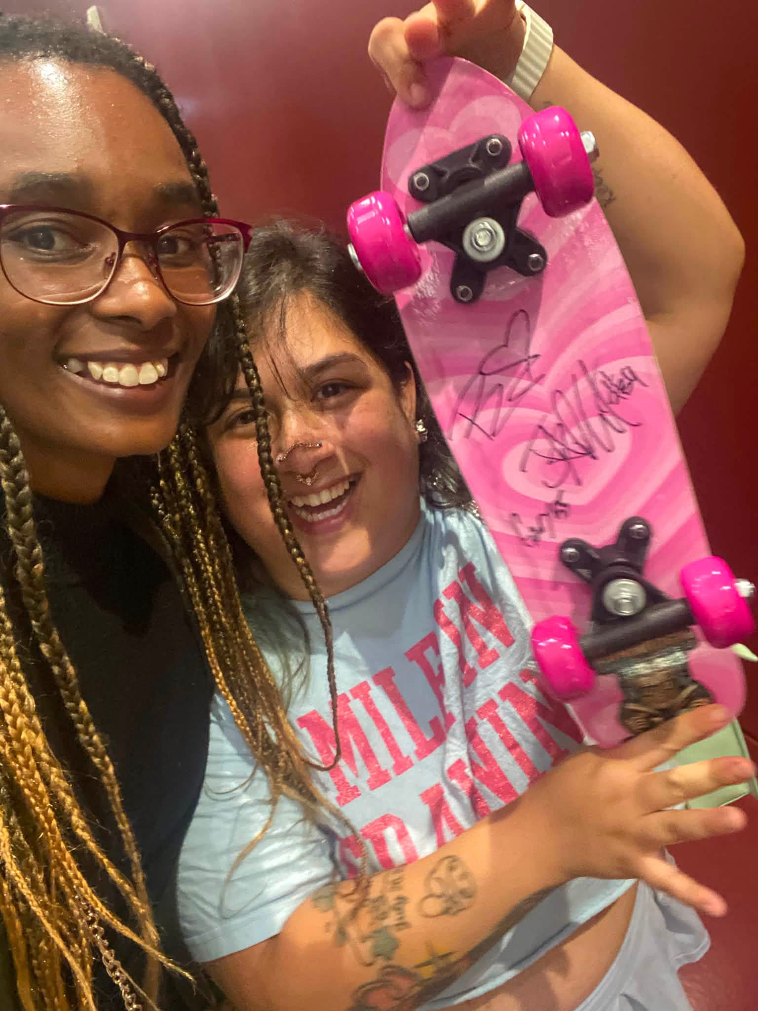 Two smiling women, one holding a pink skateboard with black wheels, in a close-up photo indoors.