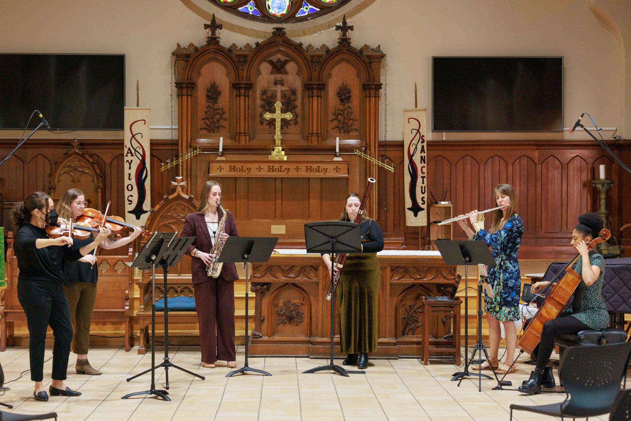 Group of musicians performing in a church with wooden altar and stained glass window, some playing instruments and others singing.