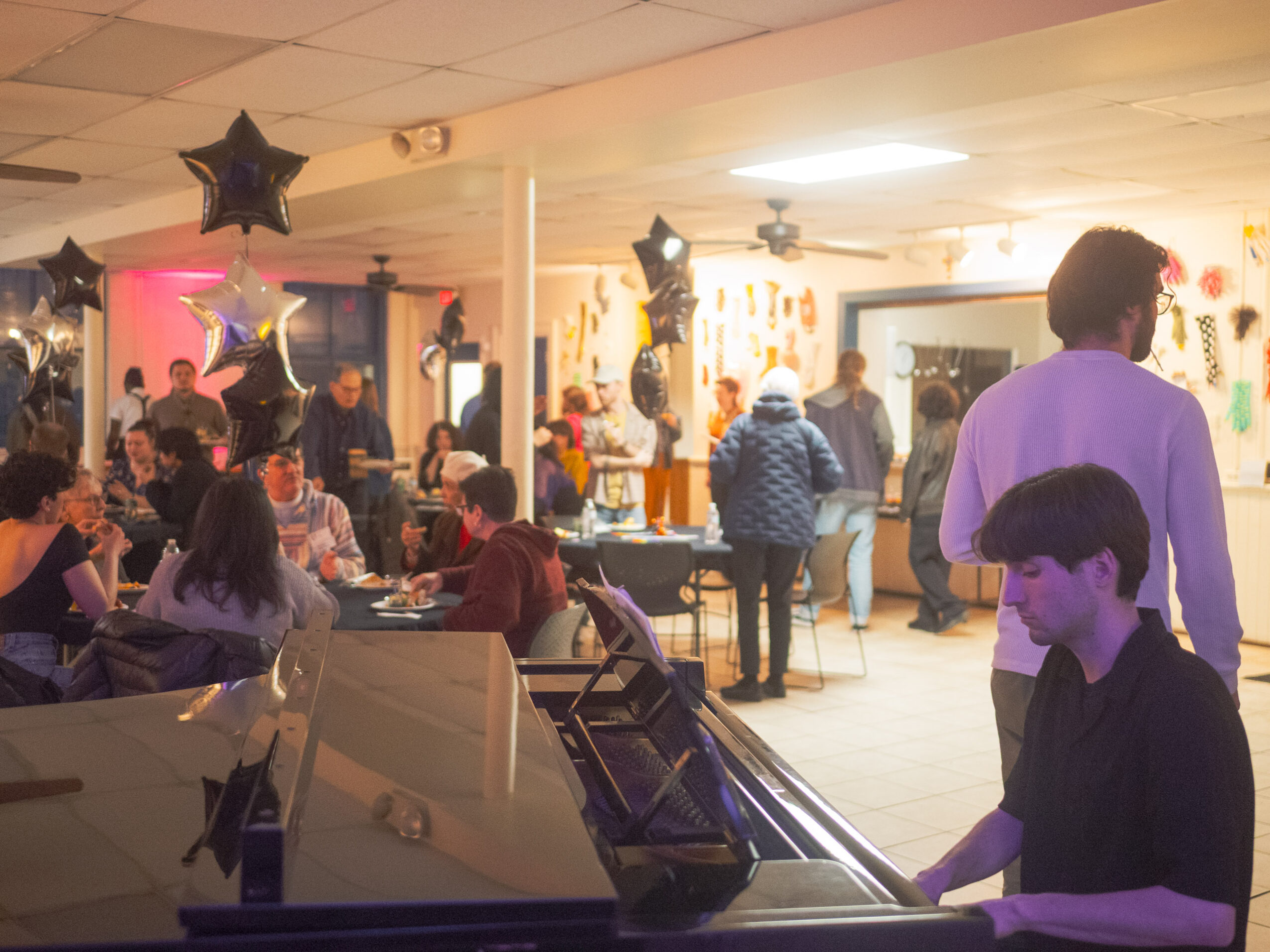 People gather in a decorated indoor space with balloons, tables, and a piano, celebrating an event or party.