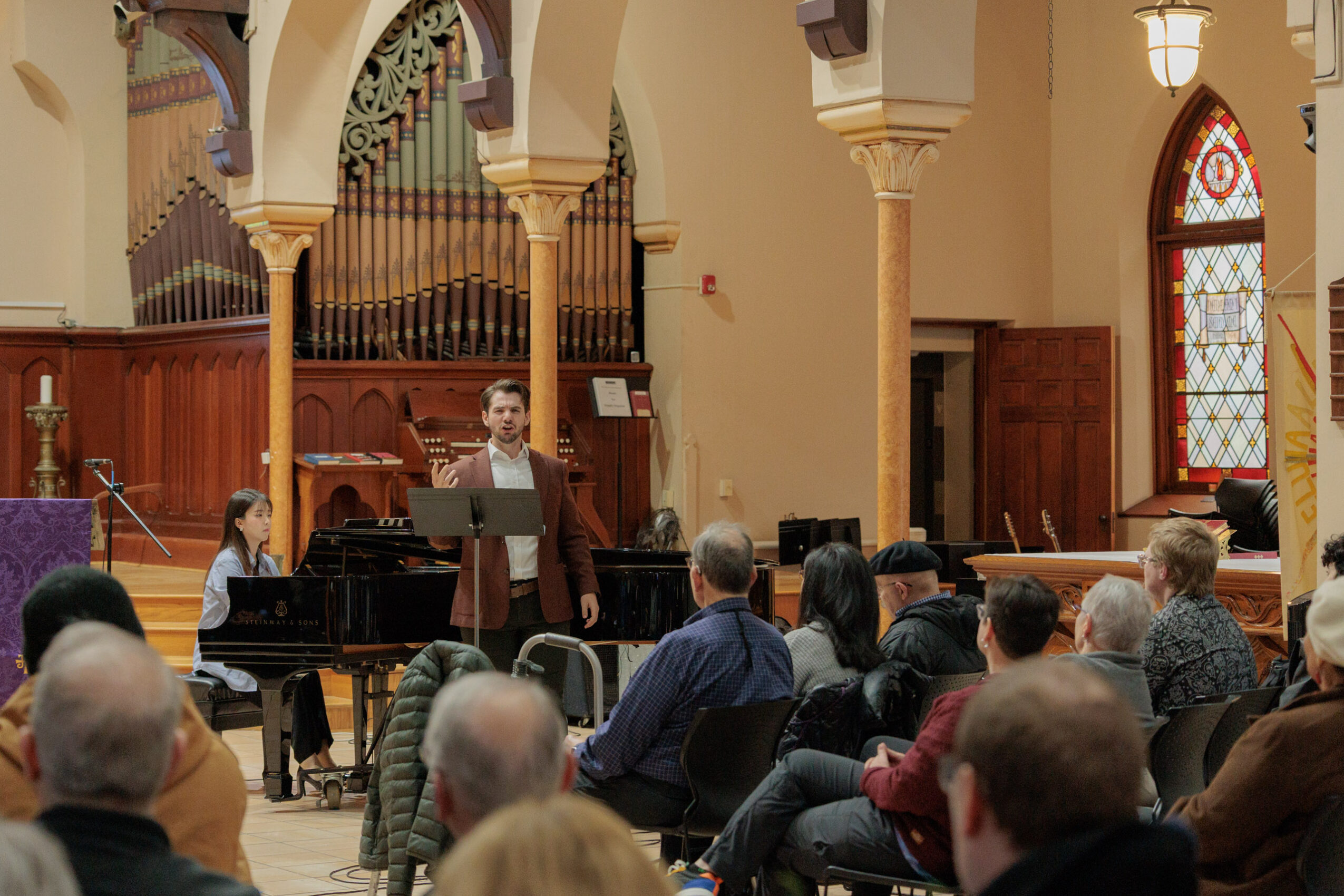 Person standing at a podium speaking to an audience inside a church with stained glass windows and wooden arches.