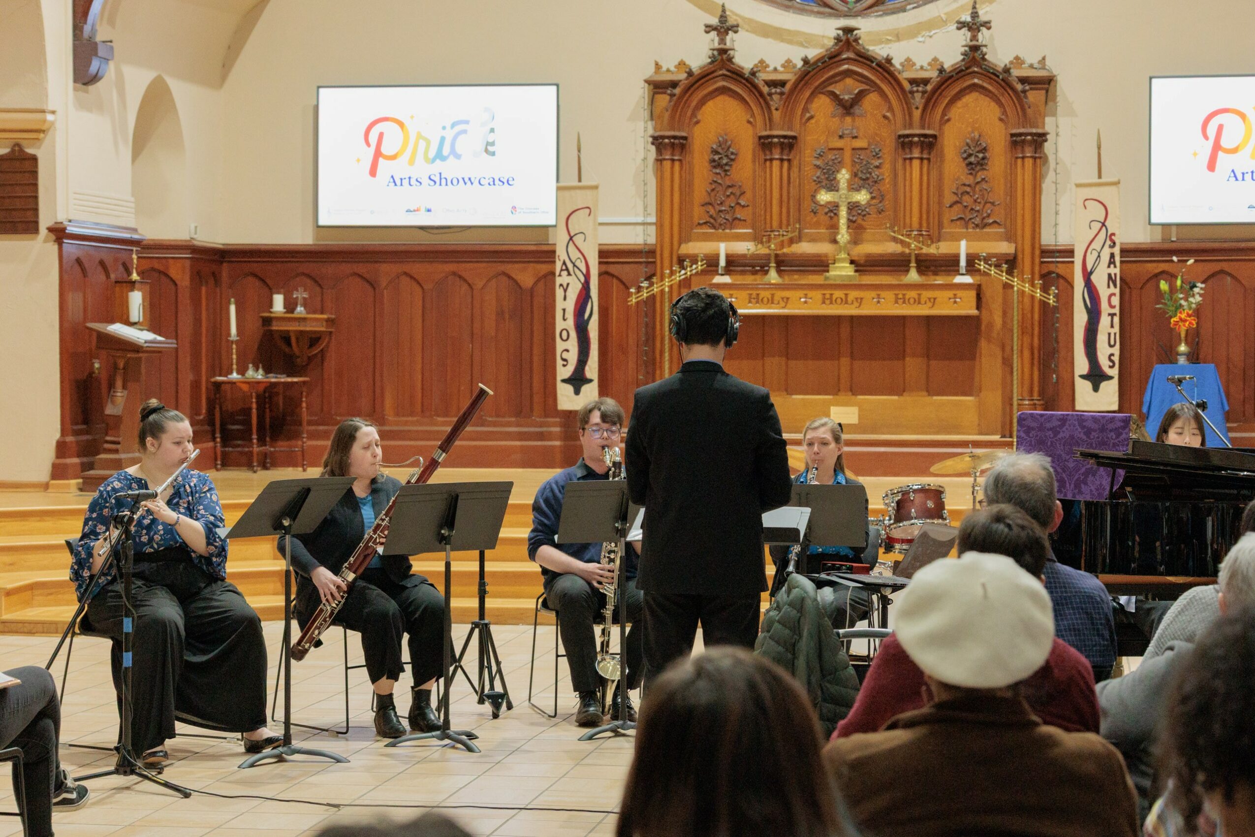 Conductor leading a small orchestra in a church with wooden altar and audience watching, two screens display a logo.