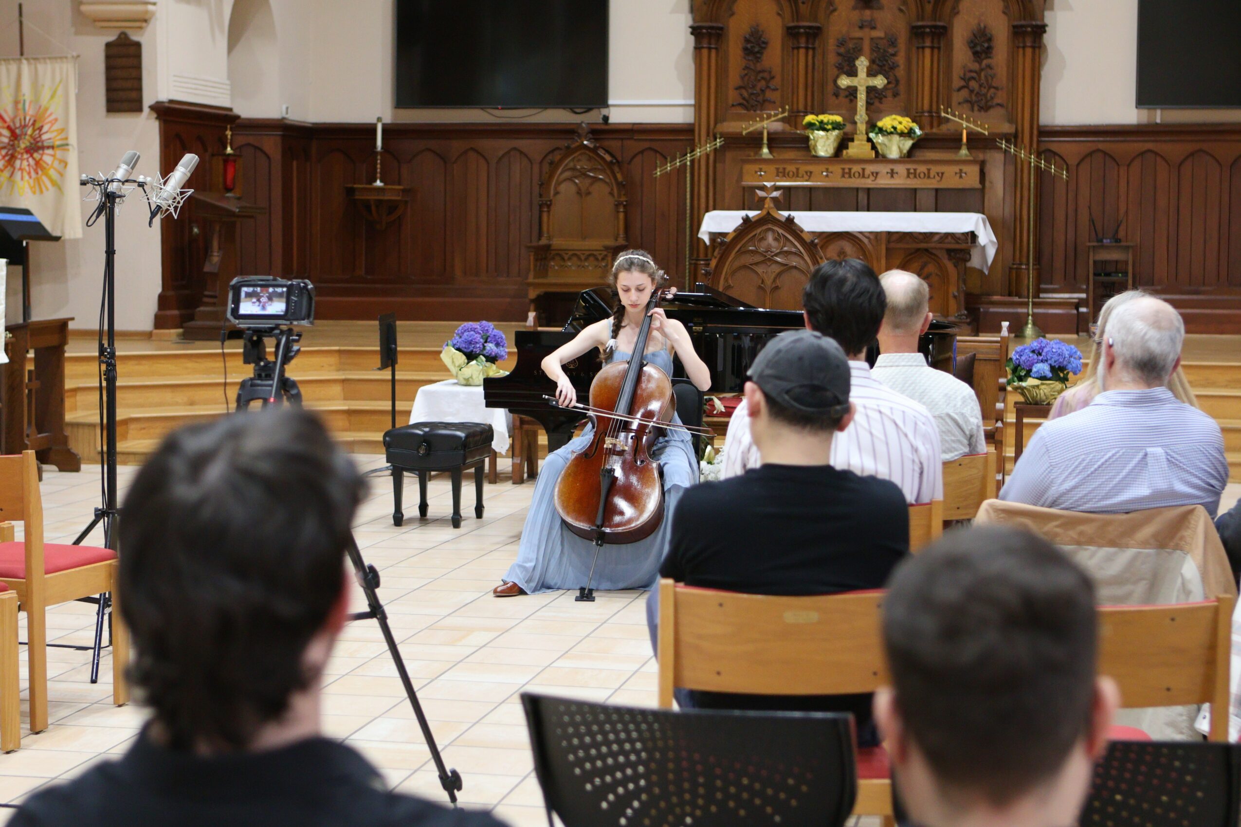 Cellist performing in a church with audience seated, wooden altar in background, flowers, and a camera on a tripod.