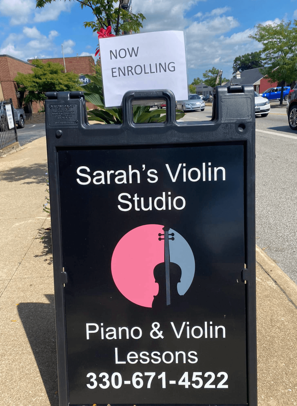 Signboard advertising Sarah's Violin Studio with contact number, outdoors on sidewalk, with a pink and blue logo, and a 'Now Enrolling' sign behind.