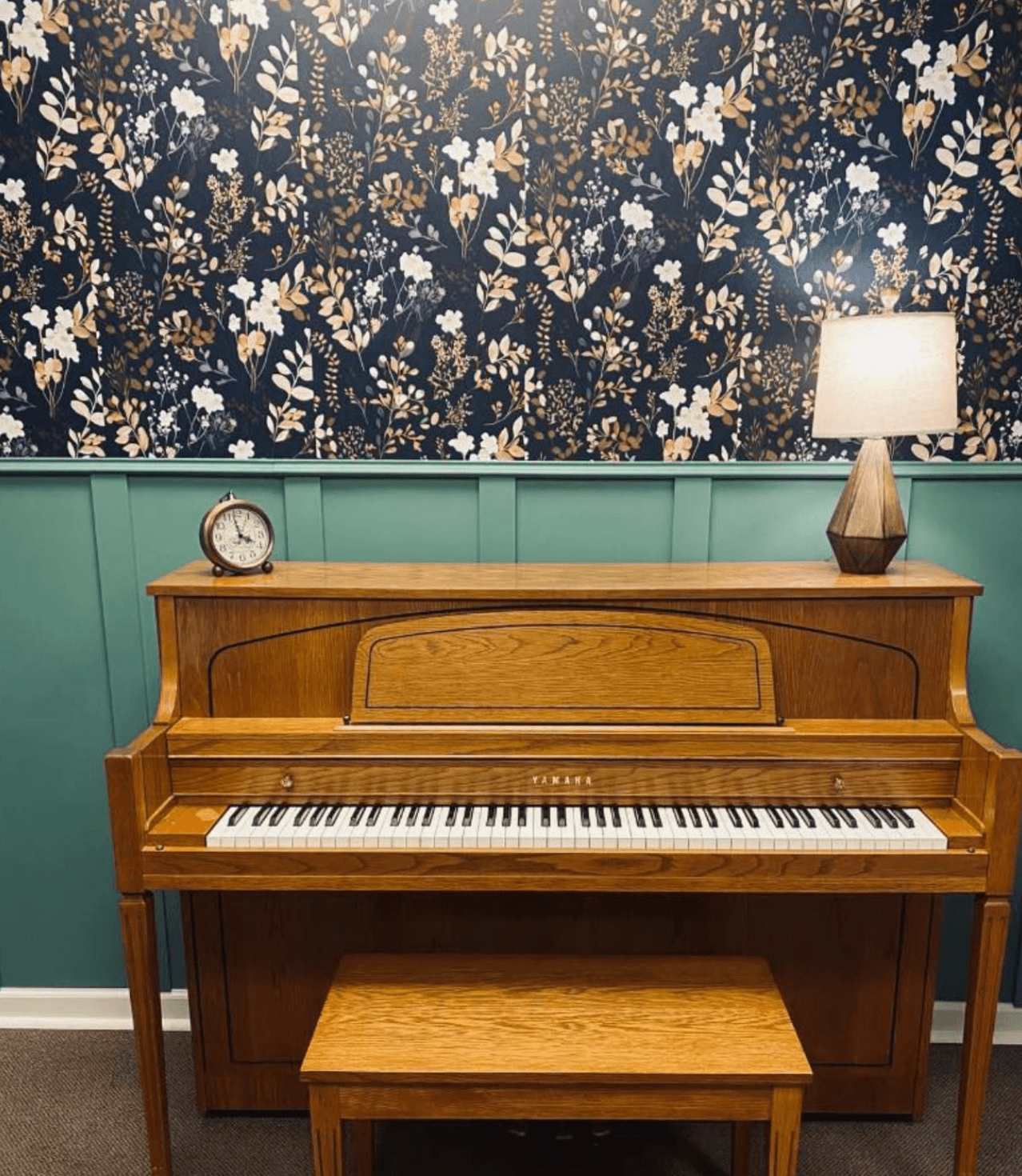 Upright wooden piano with a bench, clock, and lamp on top, against a floral wallpapered wall with teal paneling.
