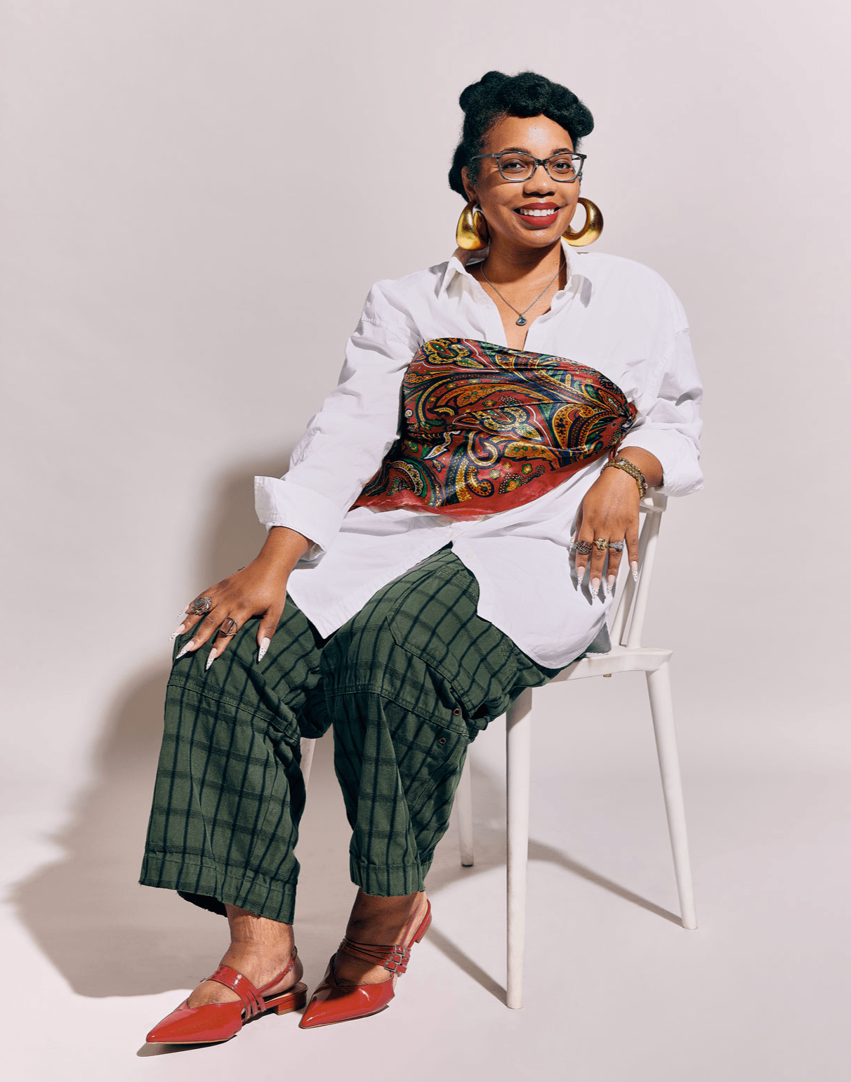 Woman sitting on a white chair, smiling, wearing a white shirt, green checkered pants, red shoes, and large earrings.