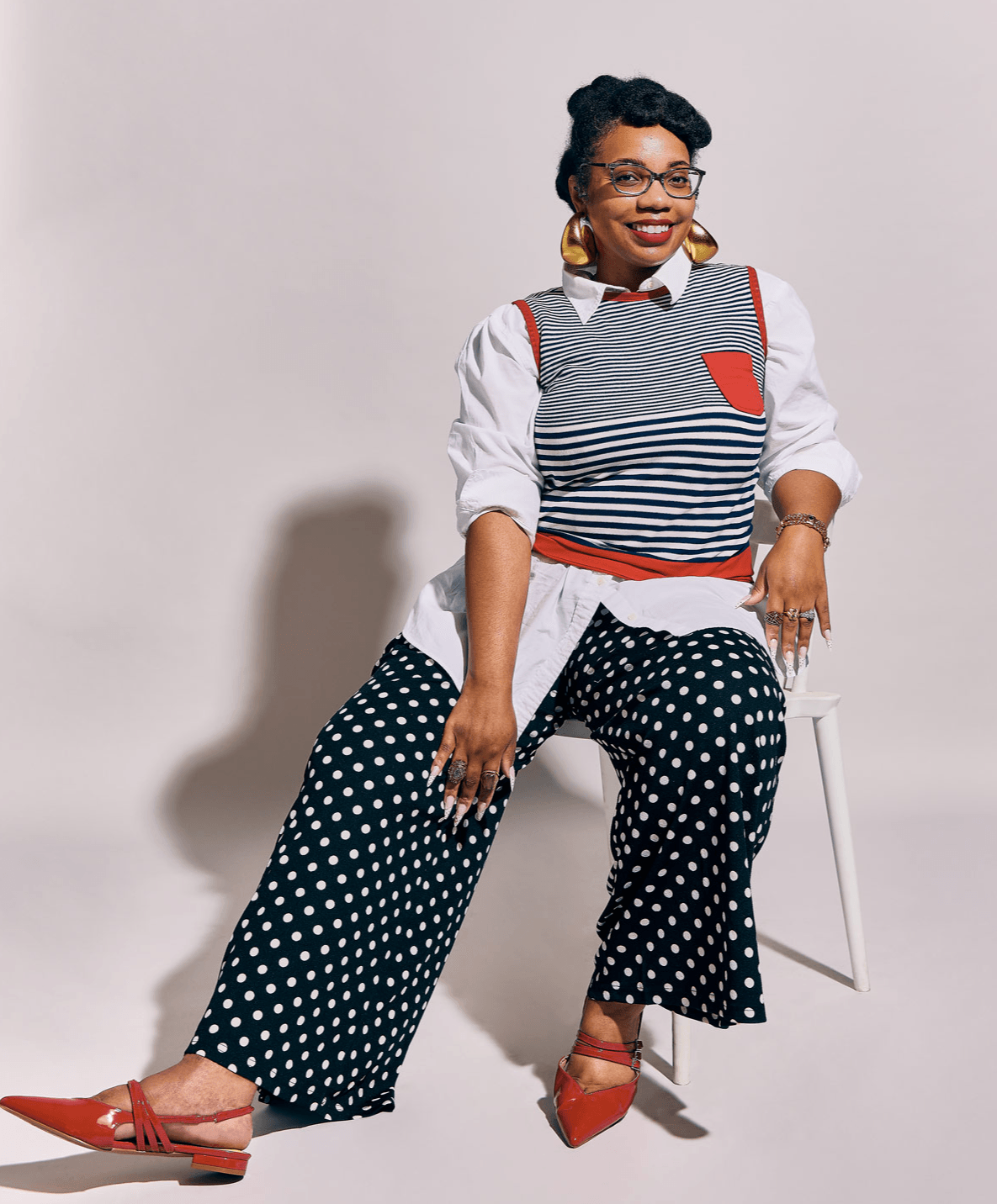 Woman sitting on a chair, smiling, wearing a layered outfit with polka dot pants and red shoes.