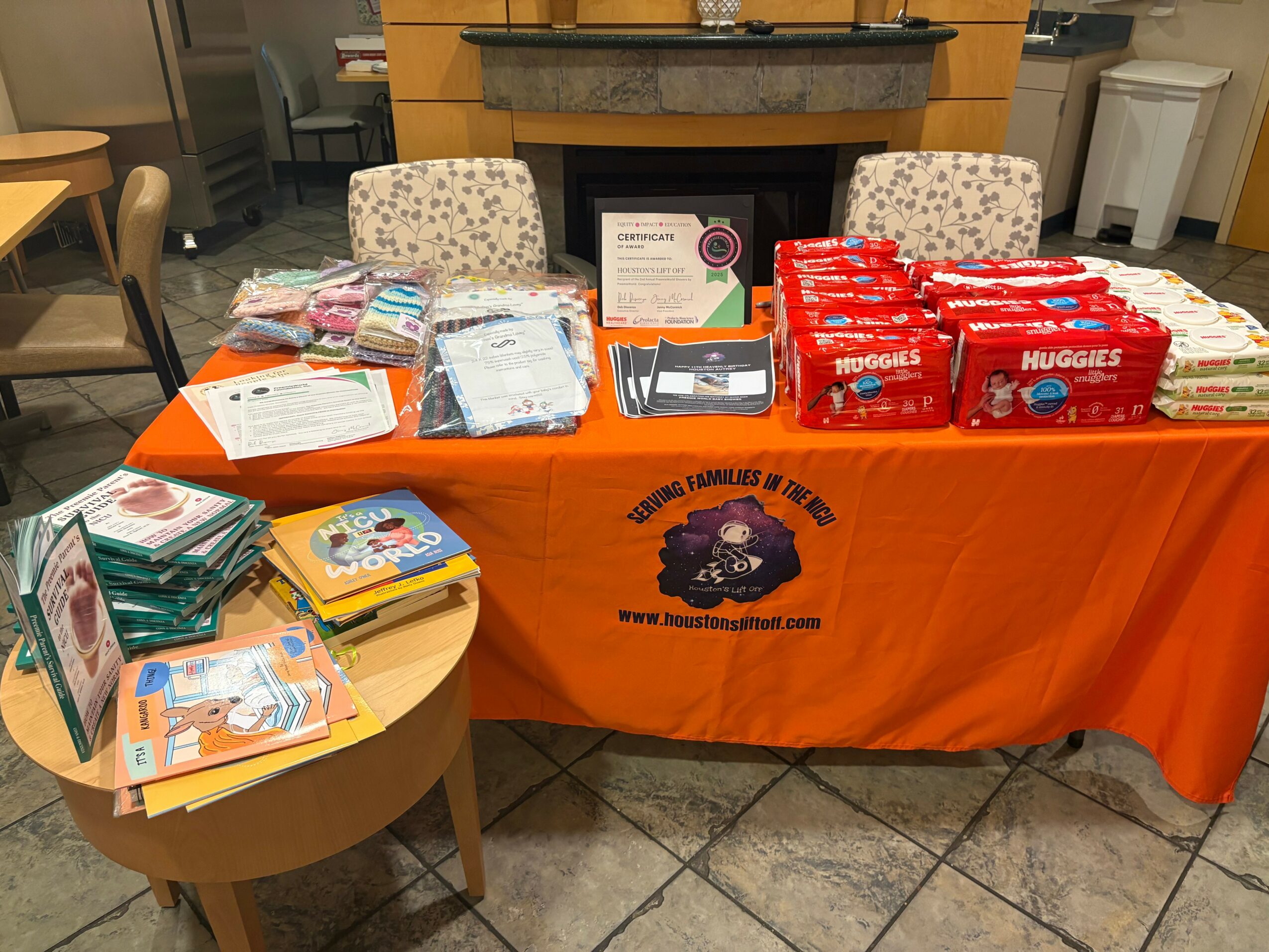 Table with diapers, supplies, and a certificate, covered with an orange cloth displaying a logo, in a room with chairs and a fireplace.