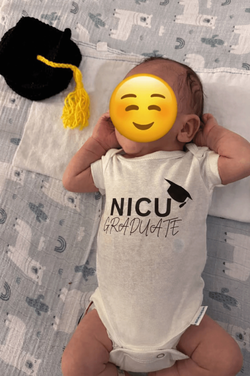 Baby lying on bed wearing a NICU graduate onesie, with a black hat with yellow tassel nearby, face covered by an emoji.