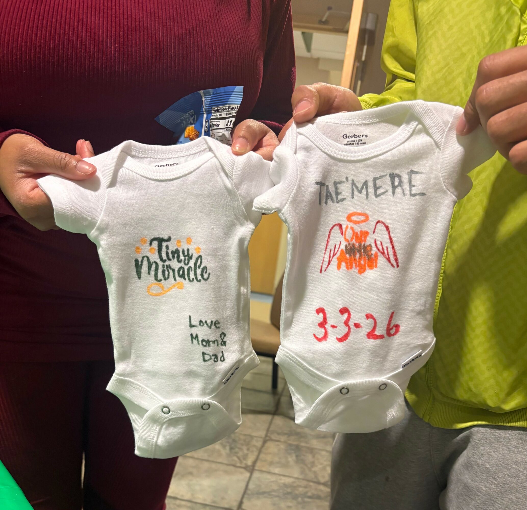Two babies' onesies held by two people, one with handwritten text and drawings, the other with printed text, in a room.