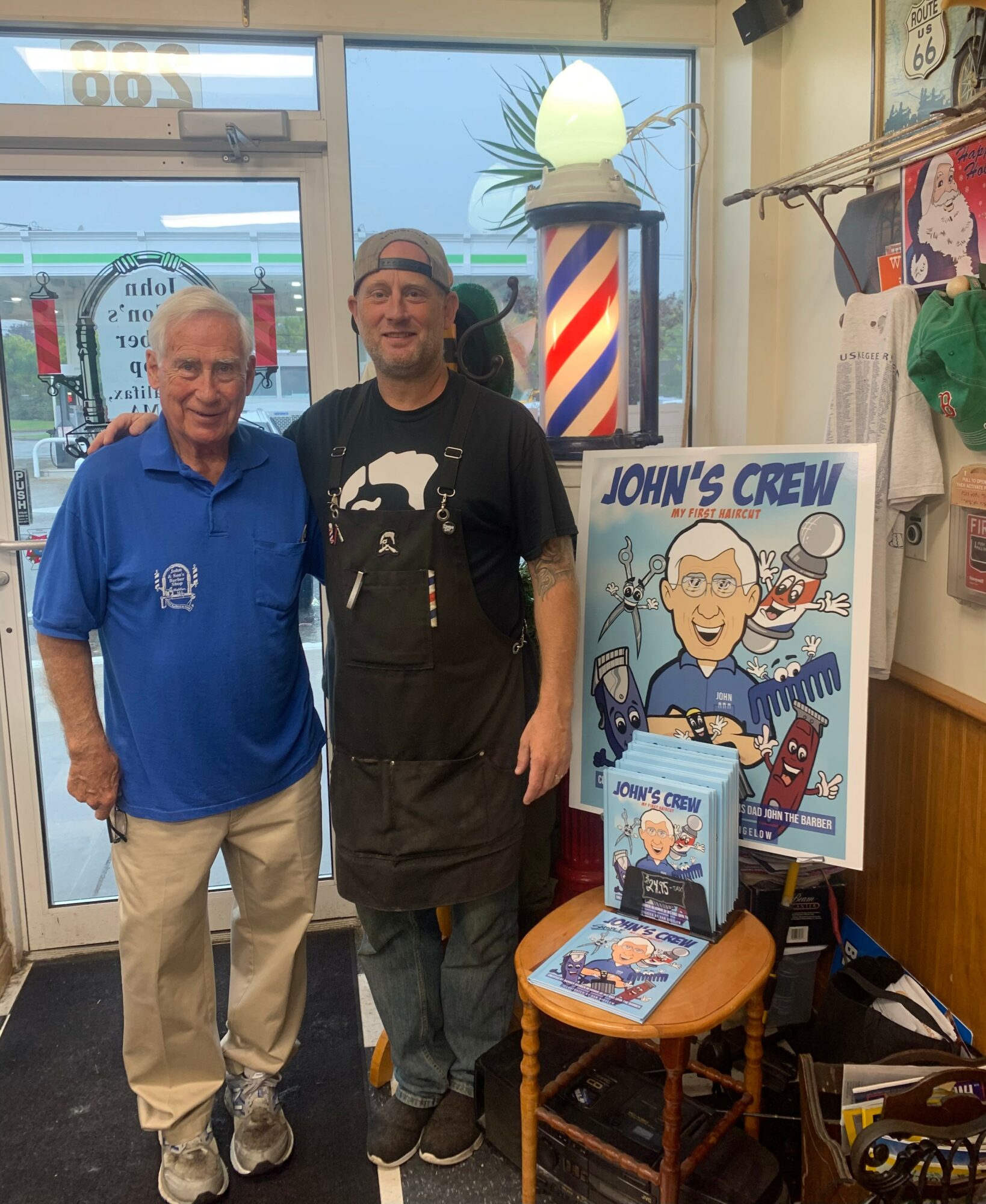 Two men standing indoors near a window, with a John’s Crew display and a chair holding books and a poster.
