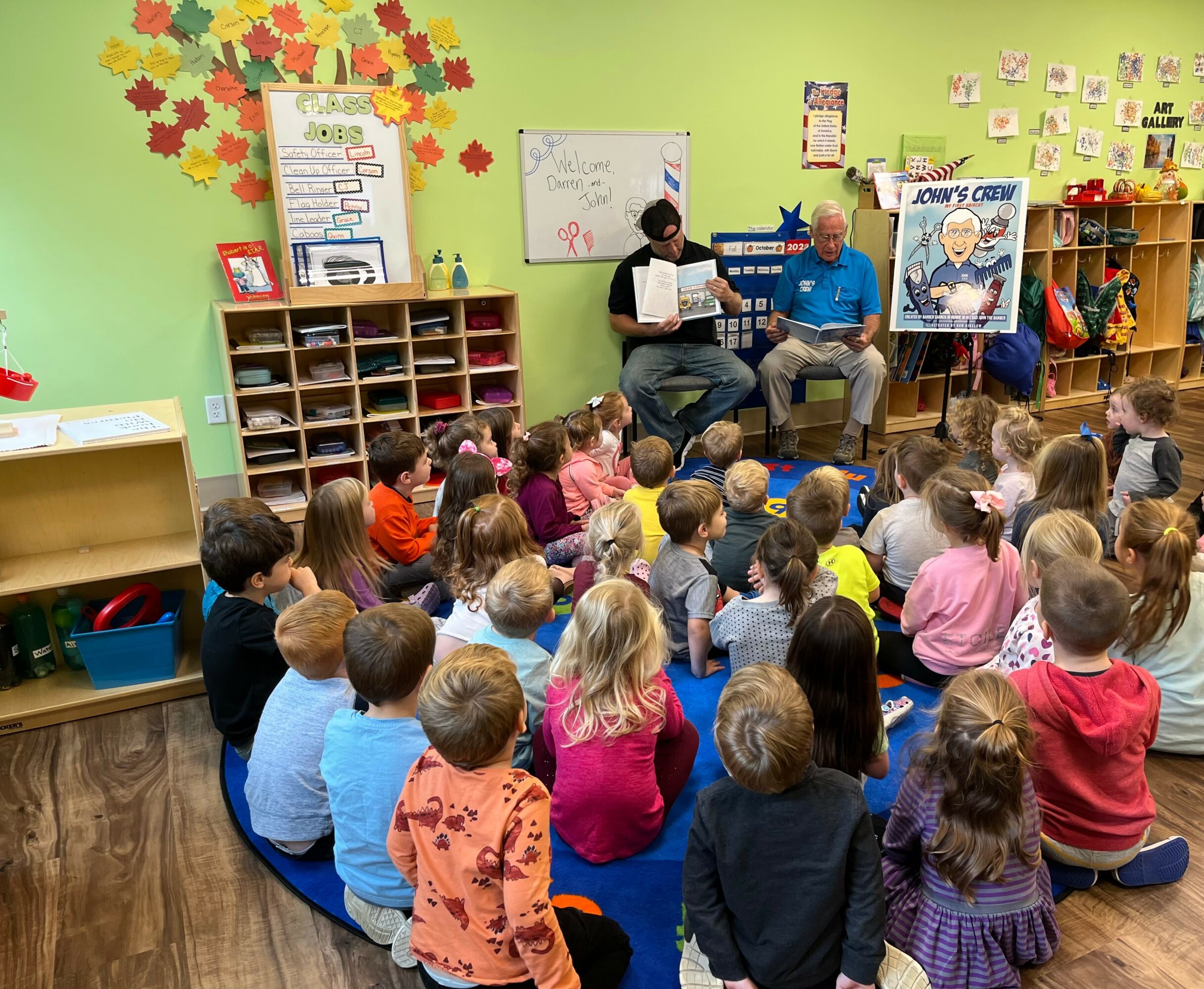 Children sit on a blue circular rug in a classroom, listening to two adults reading a book. Classroom walls are decorated with colorful posters and artwork.