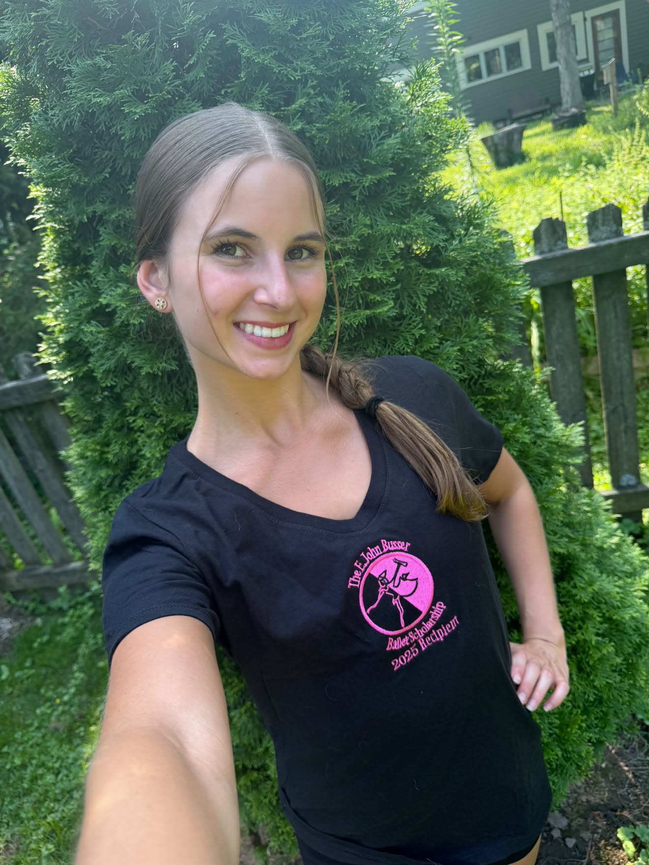 Young woman with long hair in a braid, smiling outdoors, wearing a black T-shirt with pink logo, standing near greenery.