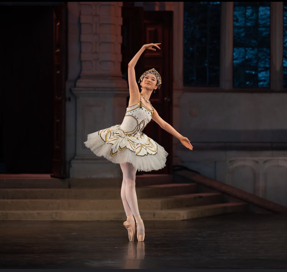 Ballet dancer in a tutu performing on stage with a building background, arms raised gracefully, standing on toes.