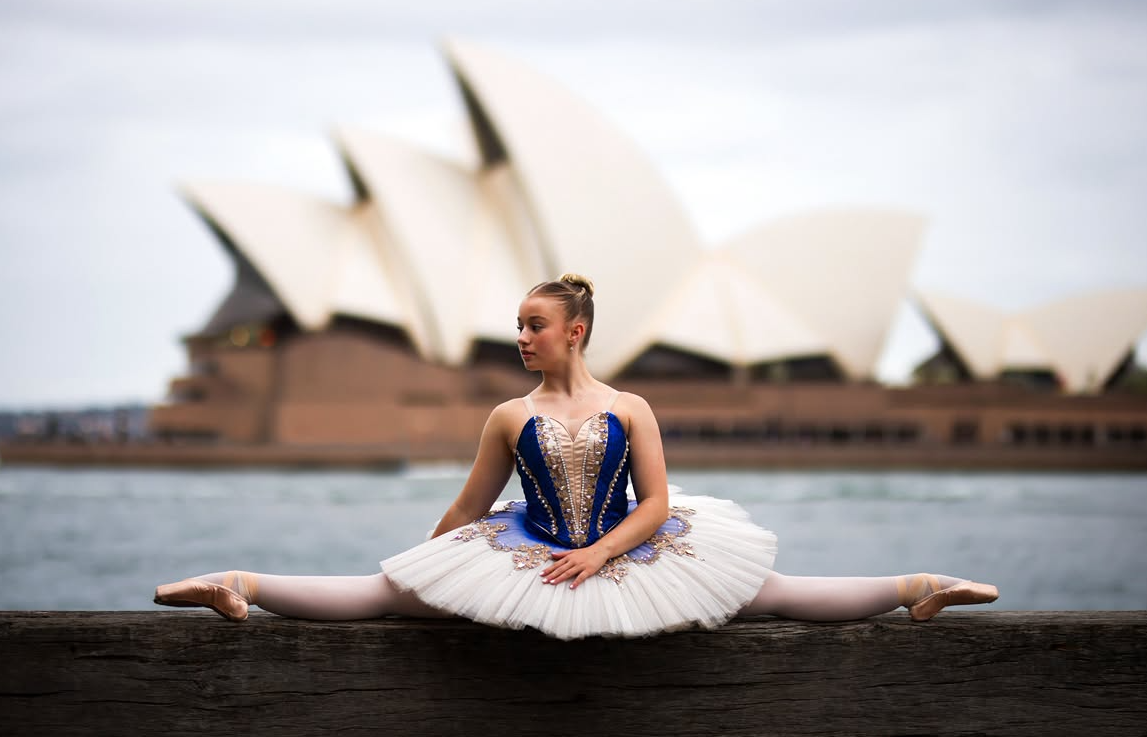 Ballet dancer in a blue and white tutu performing a split outdoors with Sydney Opera House in background.