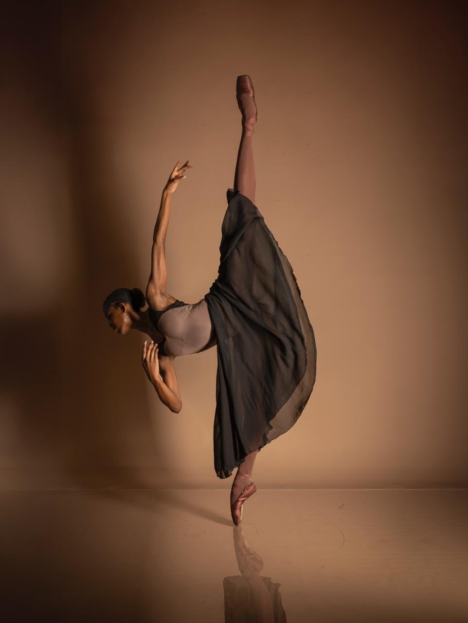Person performing a dance pose with one leg raised high and arms extended, wearing a black dress, against a brown background.