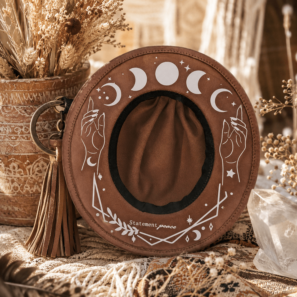 Round brown hat with moon phases and celestial designs, placed on a textured surface with dried flowers and crystals nearby.
