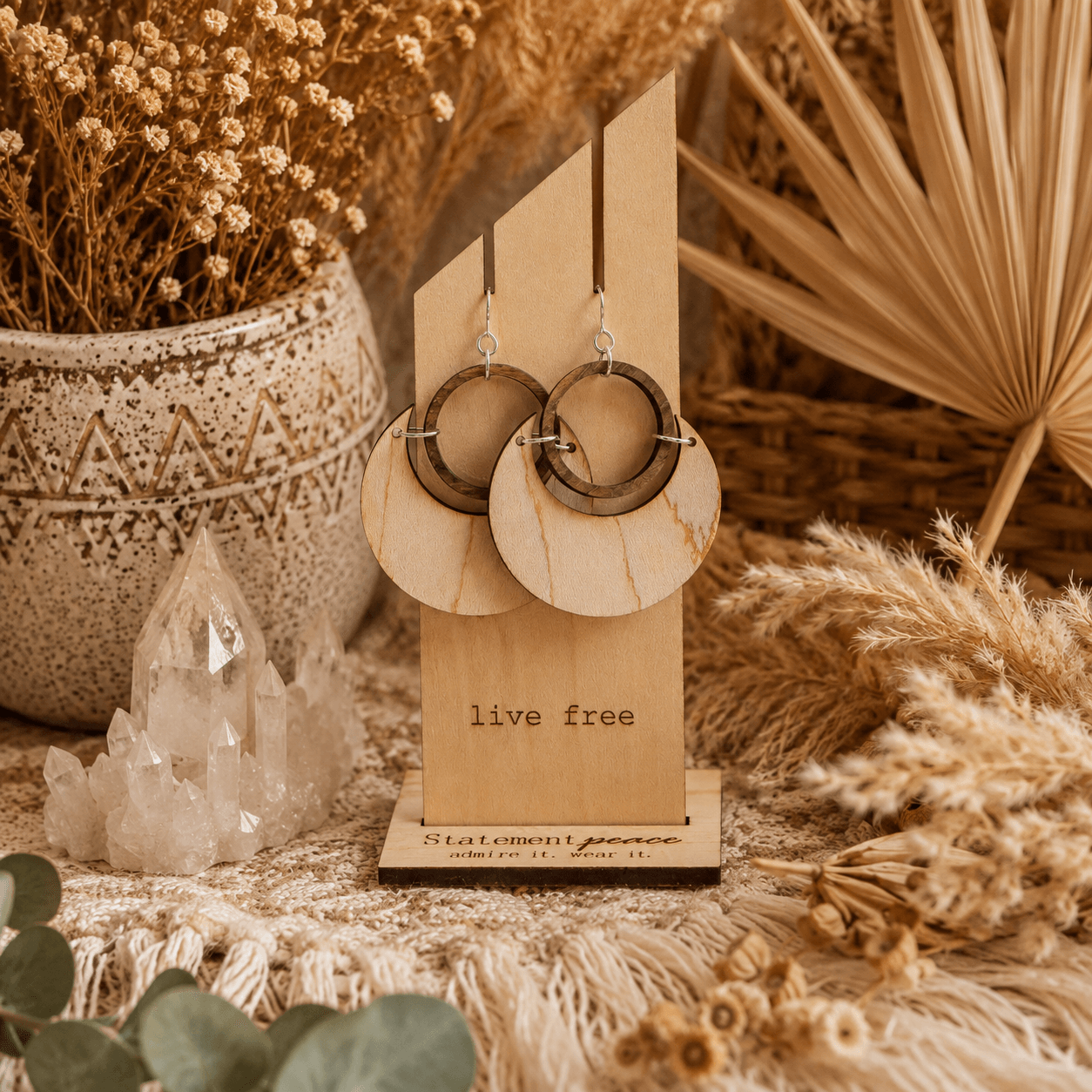 Pair of earrings with circular and semi-circular elements displayed on a wooden stand, surrounded by dried plants and crystals, with a warm, earthy aesthetic.