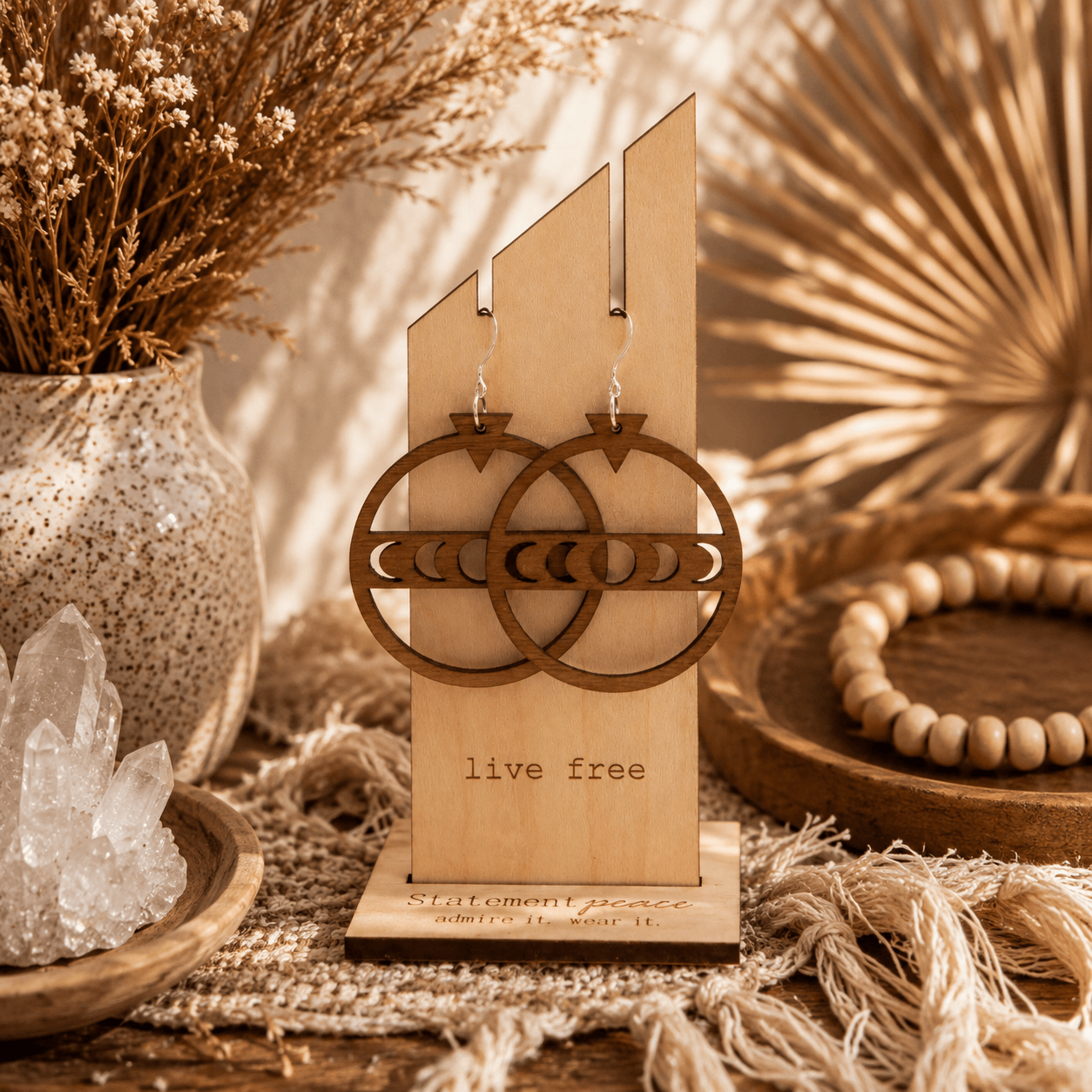 Decorative earrings hanging on a wooden display with a plant, crystals, and beads nearby, in a warm-toned setting.
