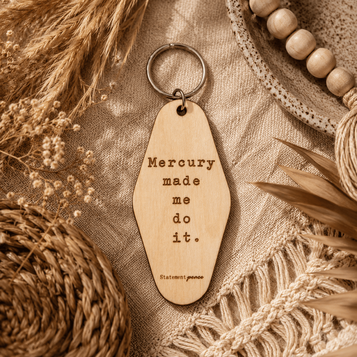 Wooden keychain with engraved text, surrounded by dried plants and woven items, on fabric background.