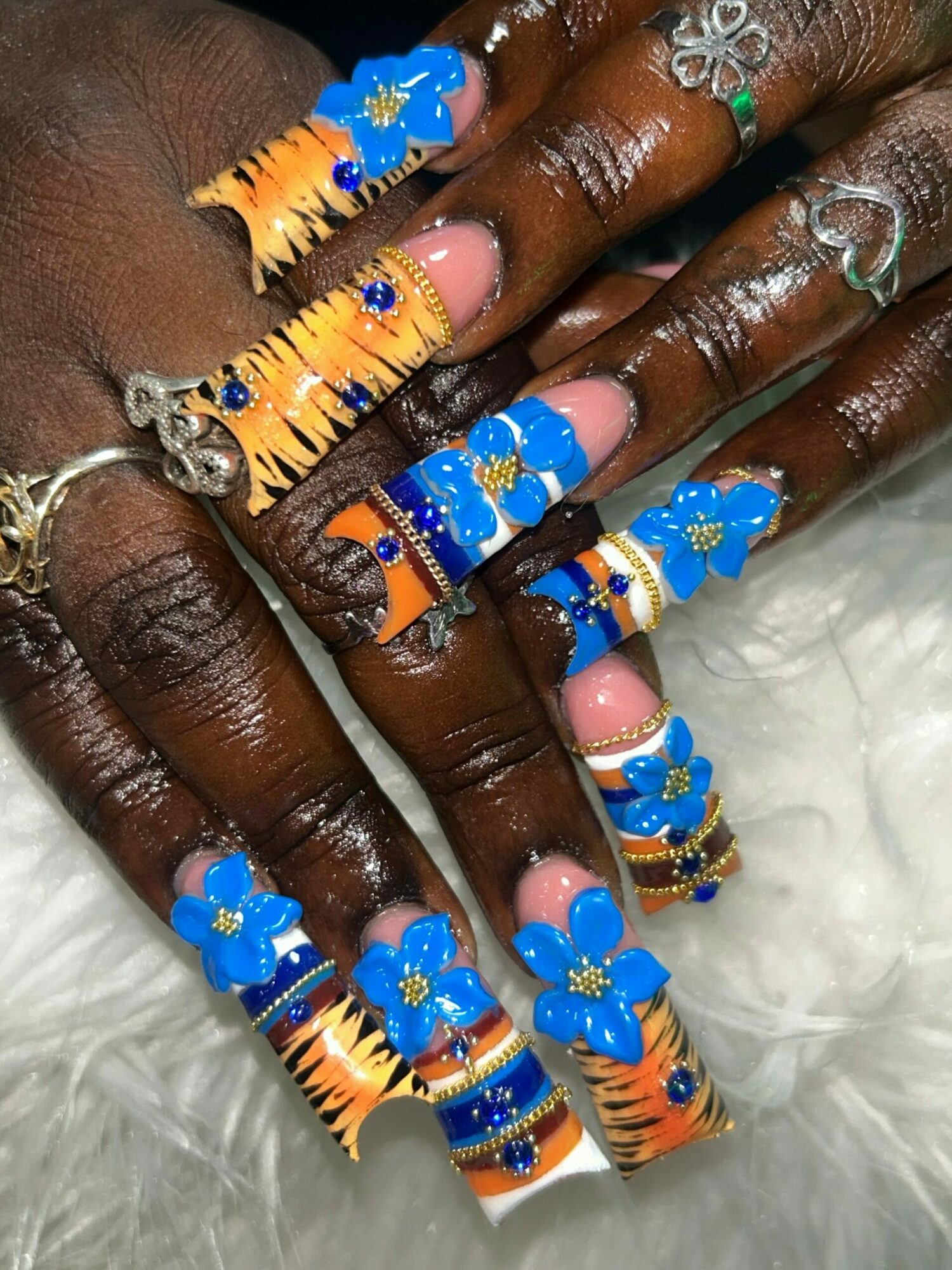 Multiple fingers with blue flower nail art and orange striped designs, adorned with rings, on a white furry background.