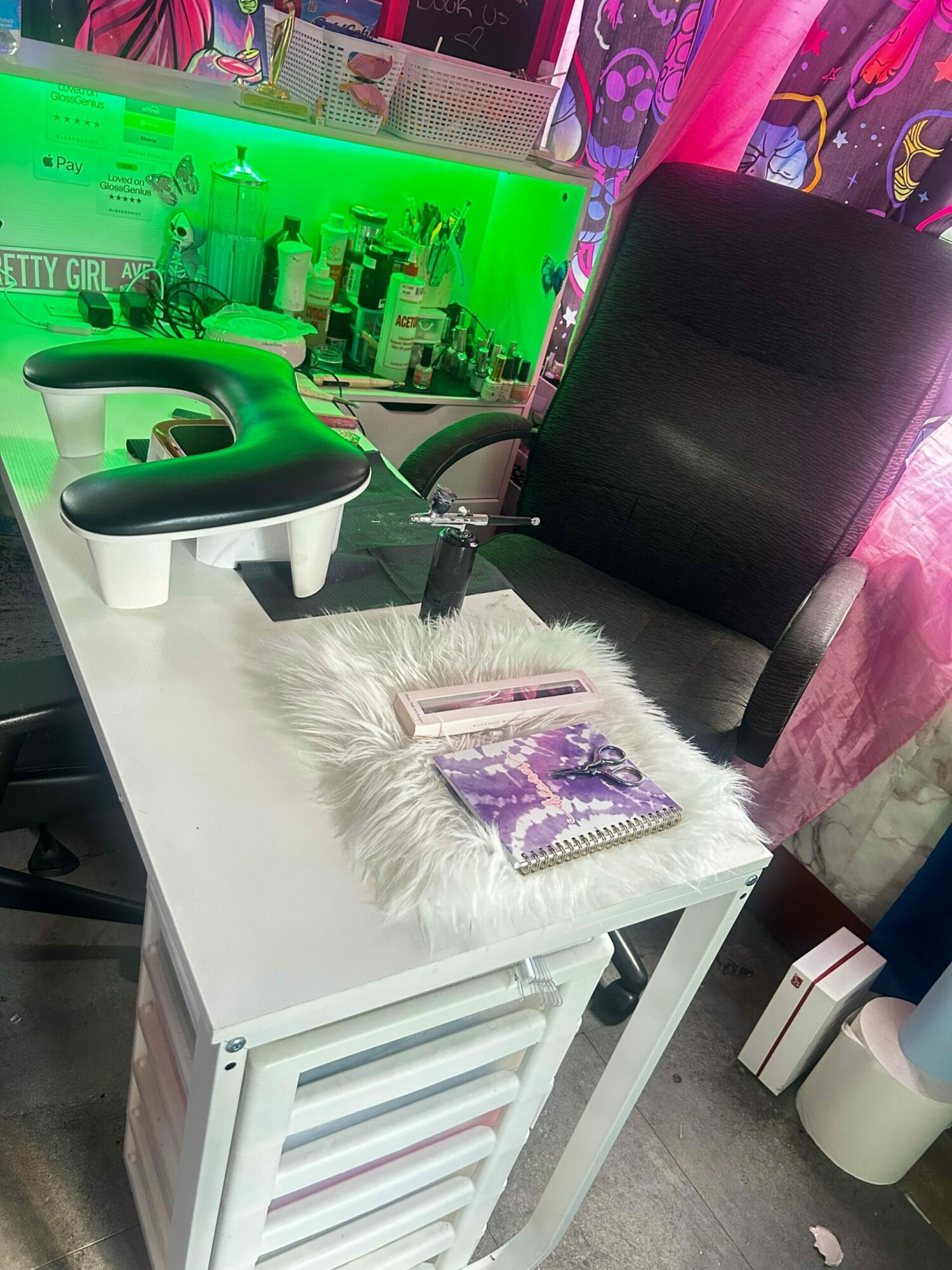 A white crib with a fluffy white rug and a purple book on top, next to a black chair and a green toy, in a room with a gray floor.
