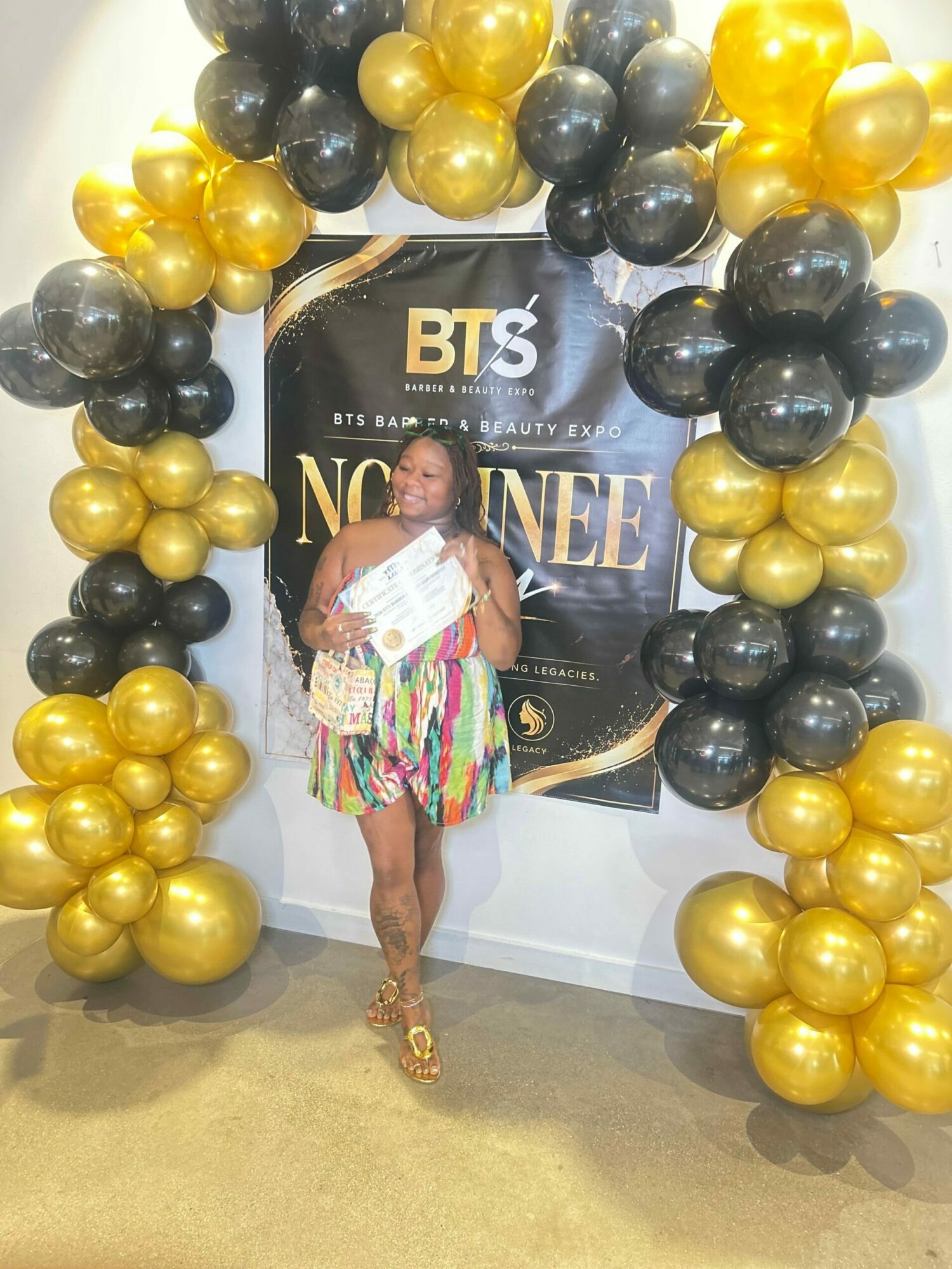 Child standing in front of a black, gold, and white backdrop with balloons, holding a certificate, at an event.