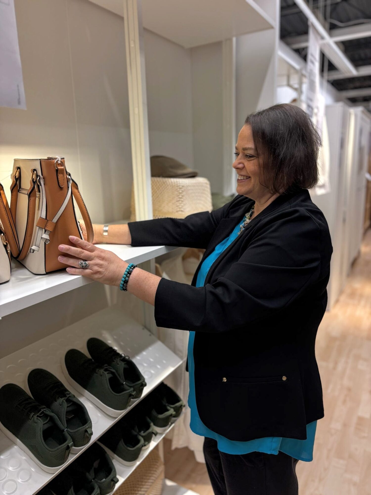 Woman with short dark hair smiling, reaching for a brown handbag on a shelf, wearing a black blazer and blue top.