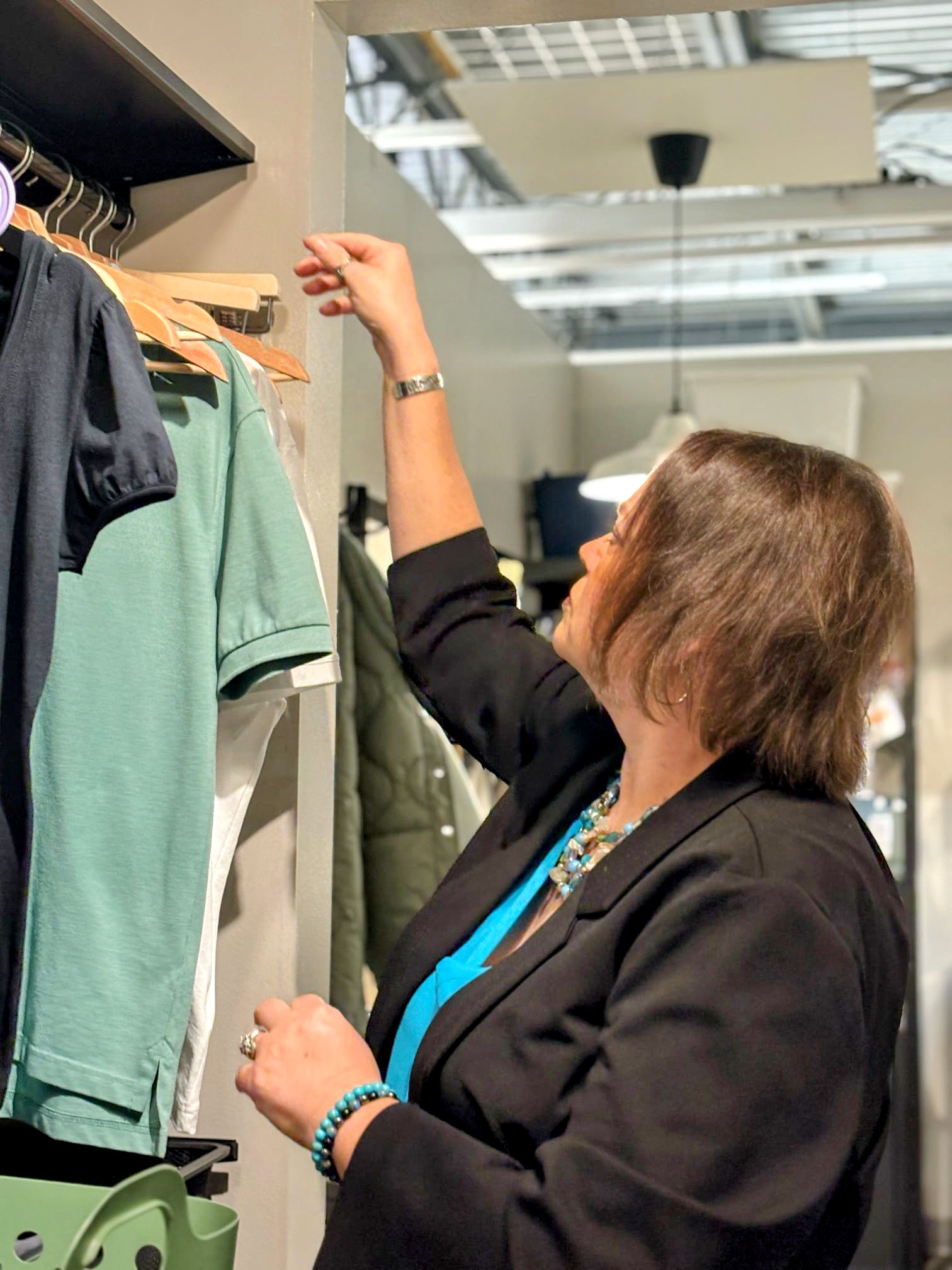 Woman shopping, reaching for a green shirt on a clothing rack in a store, with shelves and ceiling visible in background.
