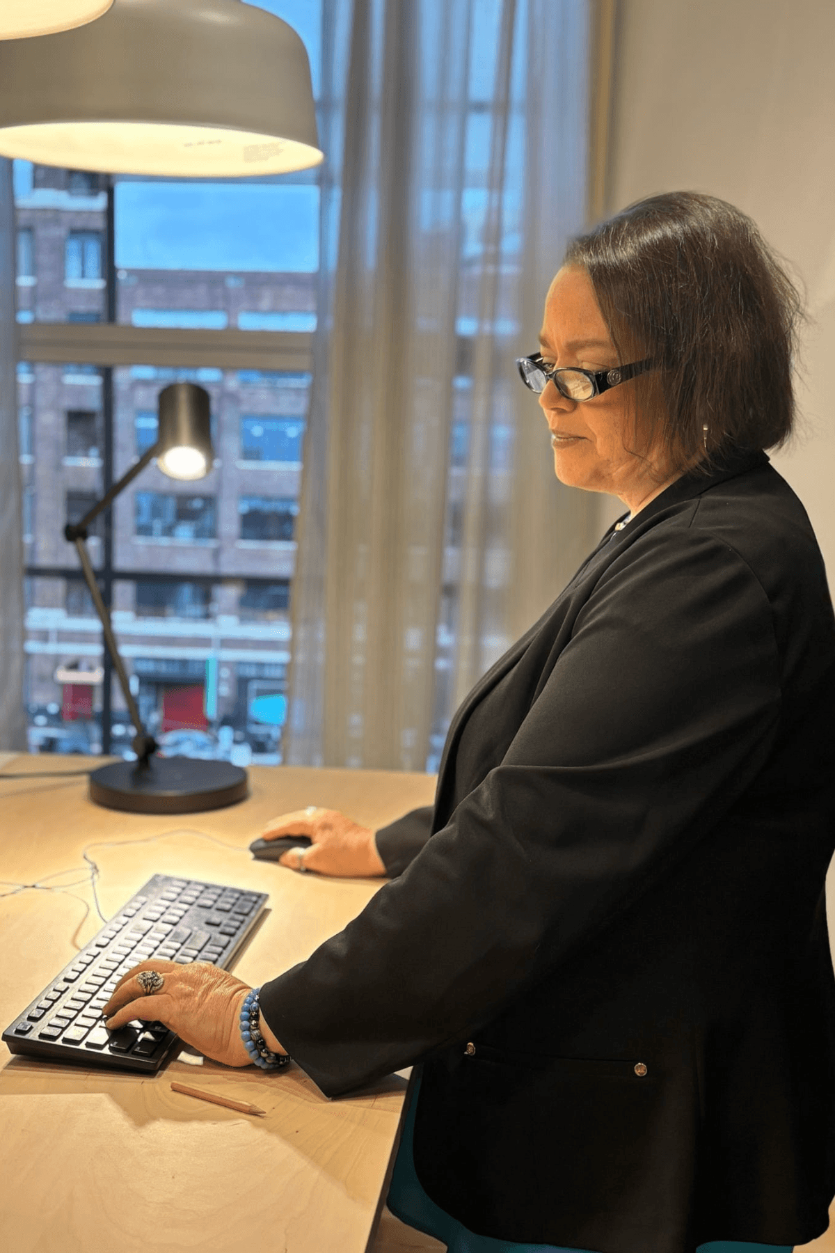 Woman with glasses in a black blazer working at a desk with a keyboard and lamp, window with buildings outside behind her.